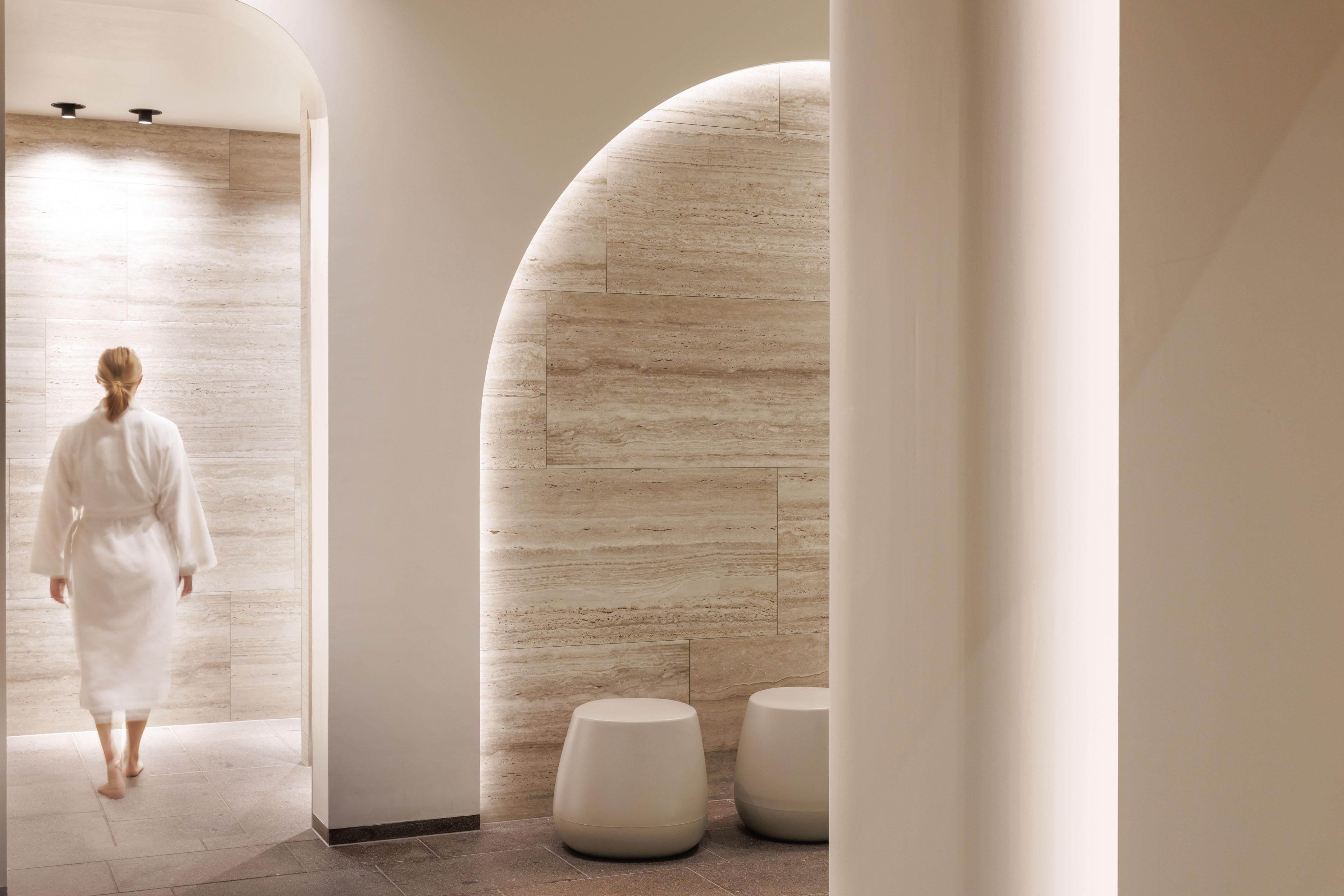 Eine Person in einem Bademantel geht in einen modernen Spa-Bereich. Die Wände sind hell und sanft beleuchtet, mit eleganten Sitzgelegenheiten.
