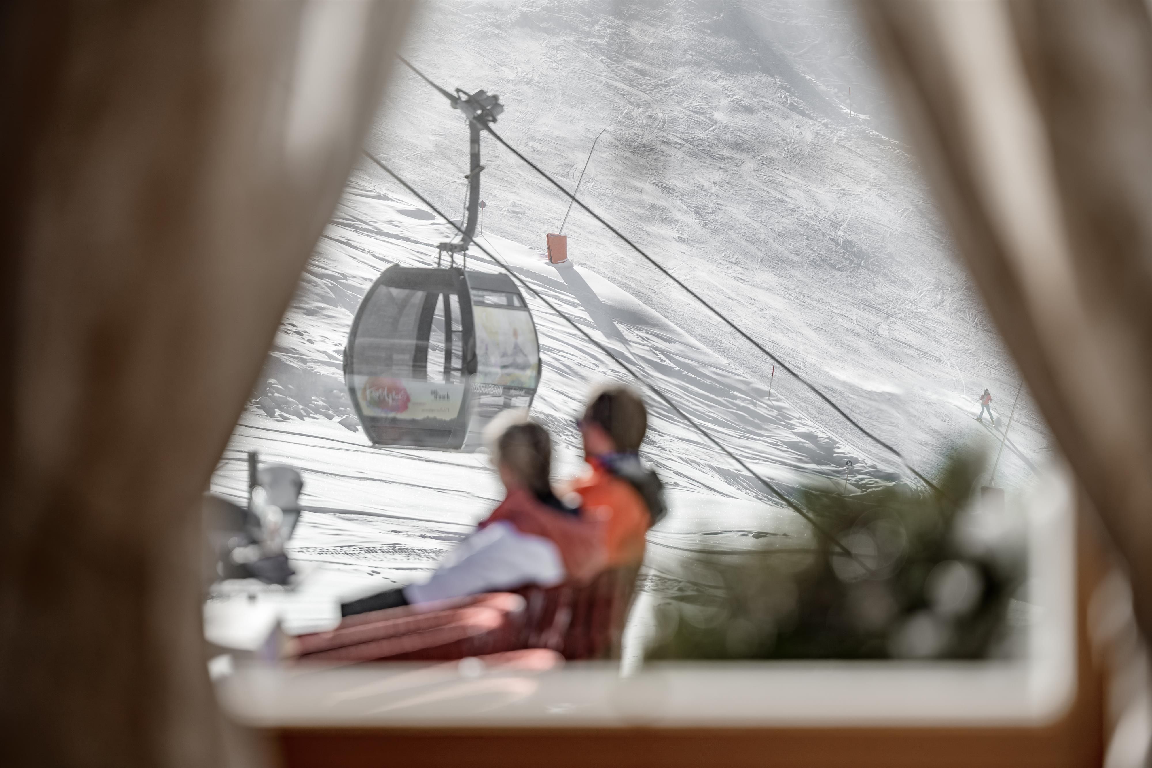 Eine gemütliche Szene mit zwei Personen, die auf einer Terrasse sitzen und die Aussicht genießen. Im Hintergrund ist eine Gondelbahn und verschneite Landschaft zu sehen.