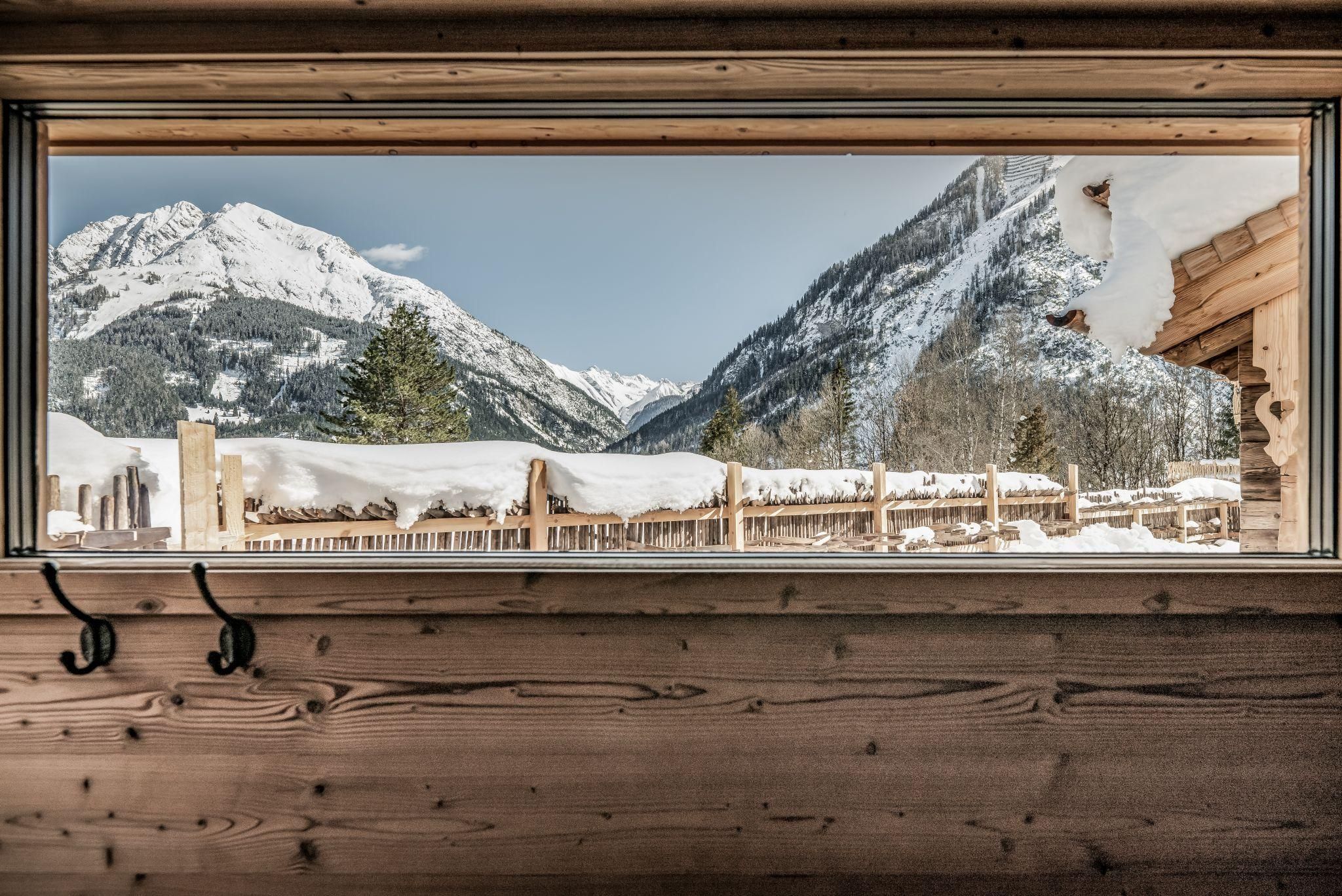 Ein Blick aus einem Holzfenster auf schneebedeckte Berge. Die Landschaft ist ruhig und winterlich.