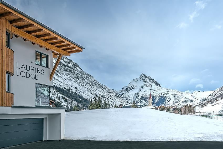 Eine moderne lodge mit dem Namen "Laurins Lodge" in einer verschneiten Berglandschaft. Im Hintergrund sind majestätische Berge unter einem bewölkten Himmel zu sehen.