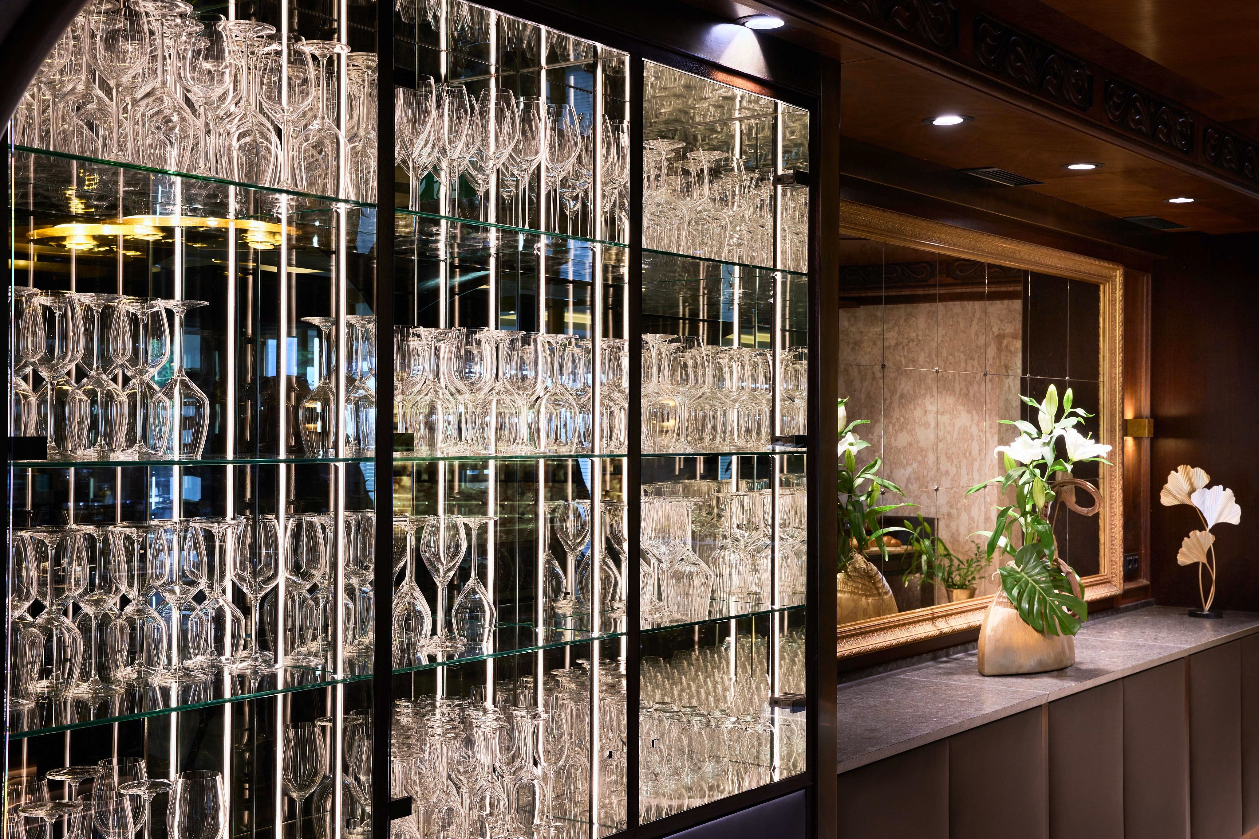 Ein elegantes Glasregal mit vielen Gläsern in einer stilvollen Bar. Im Hintergrund sind Pflanzen und eine gemütliche Einrichtung zu sehen.