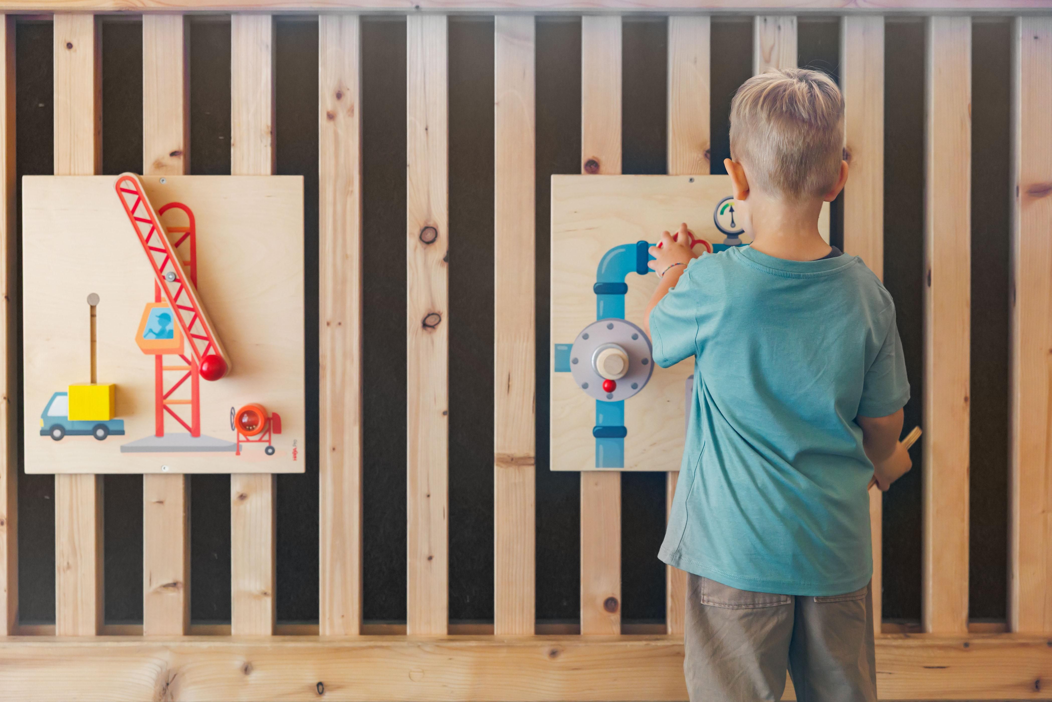 Ein Junge steht vor zwei Holztafeln mit bunten Baustellenmotiven. Er interagiert mit einem Spielzeugkran und einer Wasserleitung.