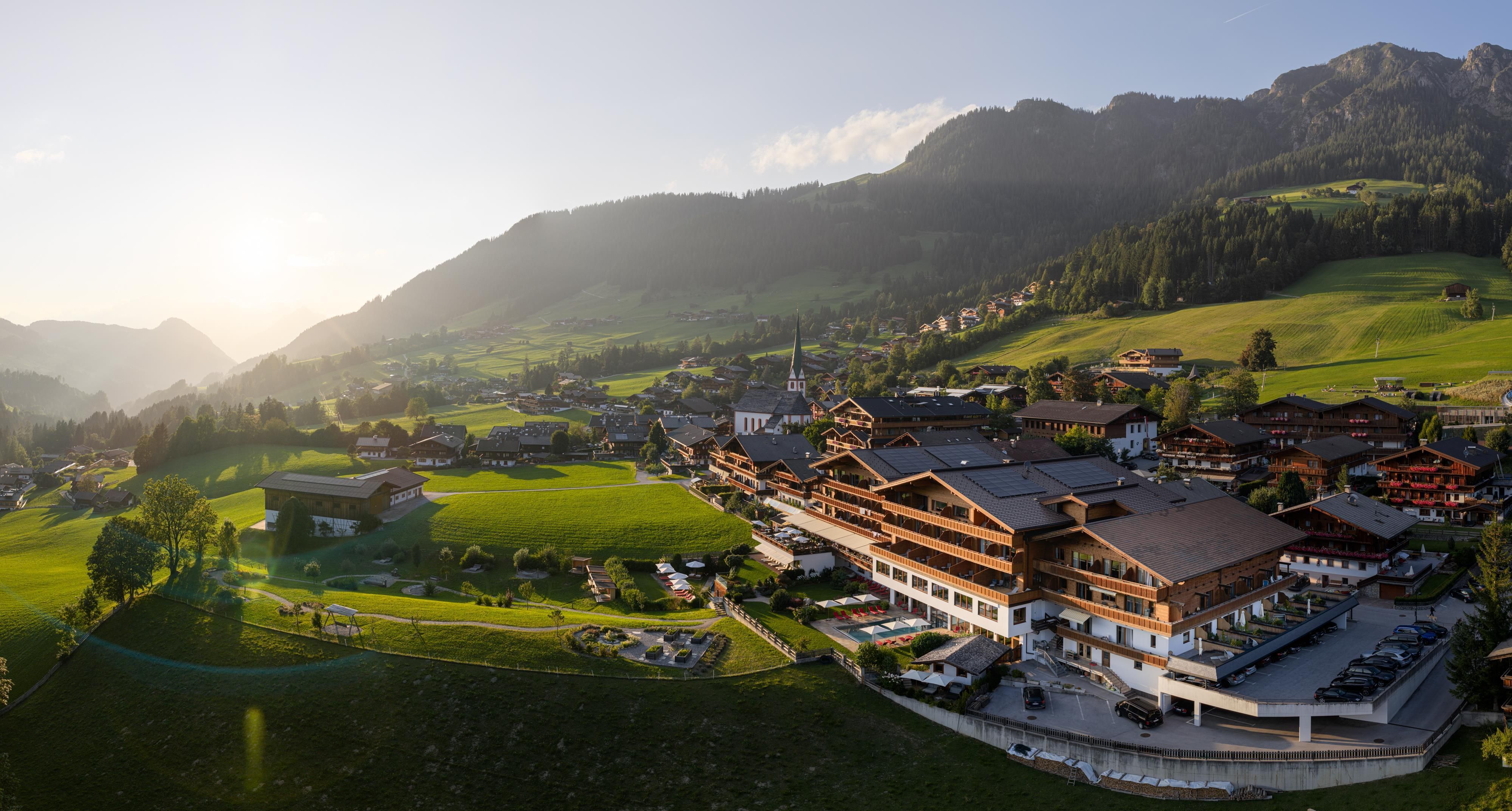 Eine malerische Landschaft mit grünen Wiesen und Bergen im Hintergrund. Im Vordergrund befindet sich ein charmantes Dorf mit traditionellen Gebäuden.