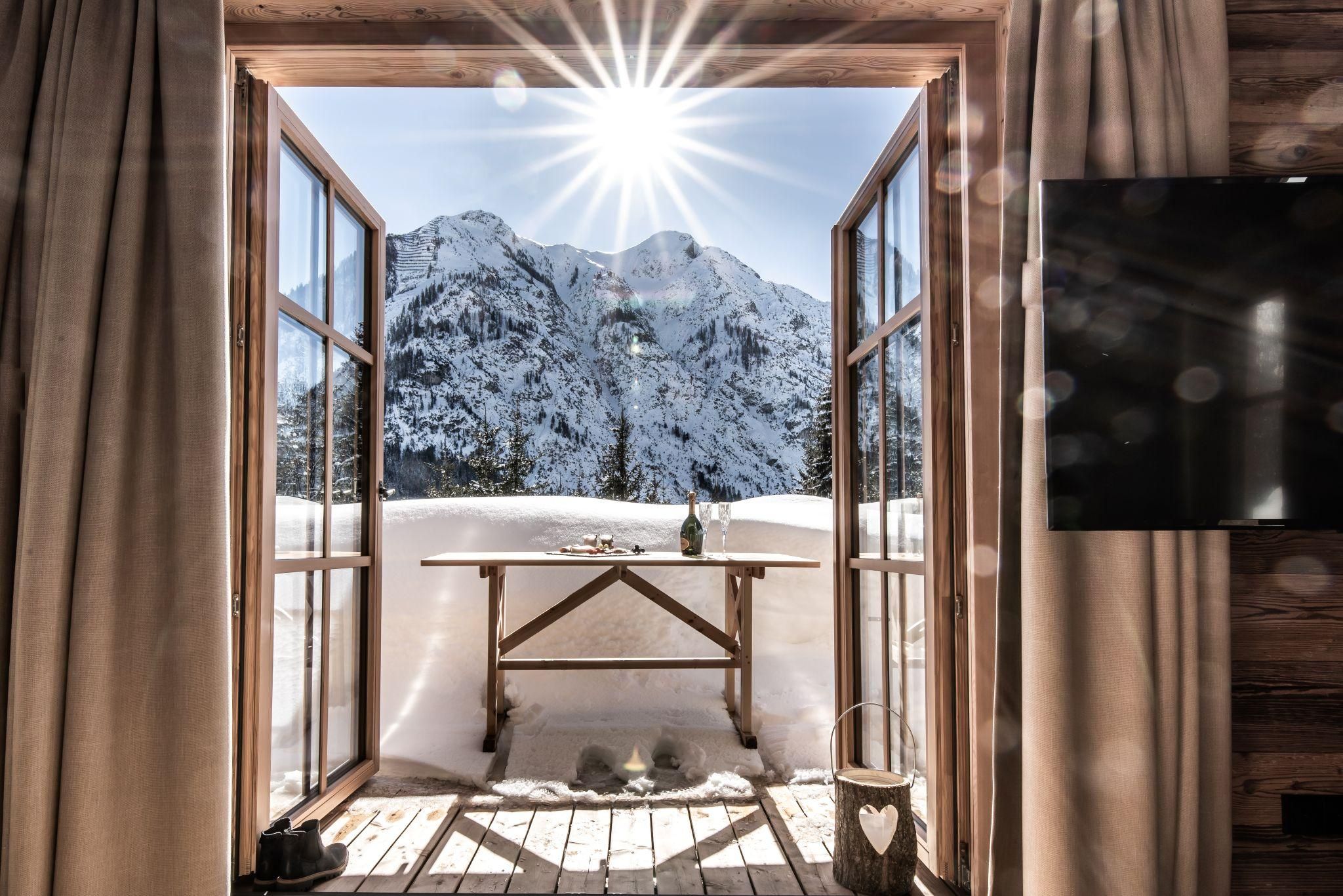 Ein Blick aus einem Holzfenster auf beeindruckende schneebedeckte Berge und strahlenden Sonnenschein. Draußen steht ein rustikaler Tisch mit einer Flasche.