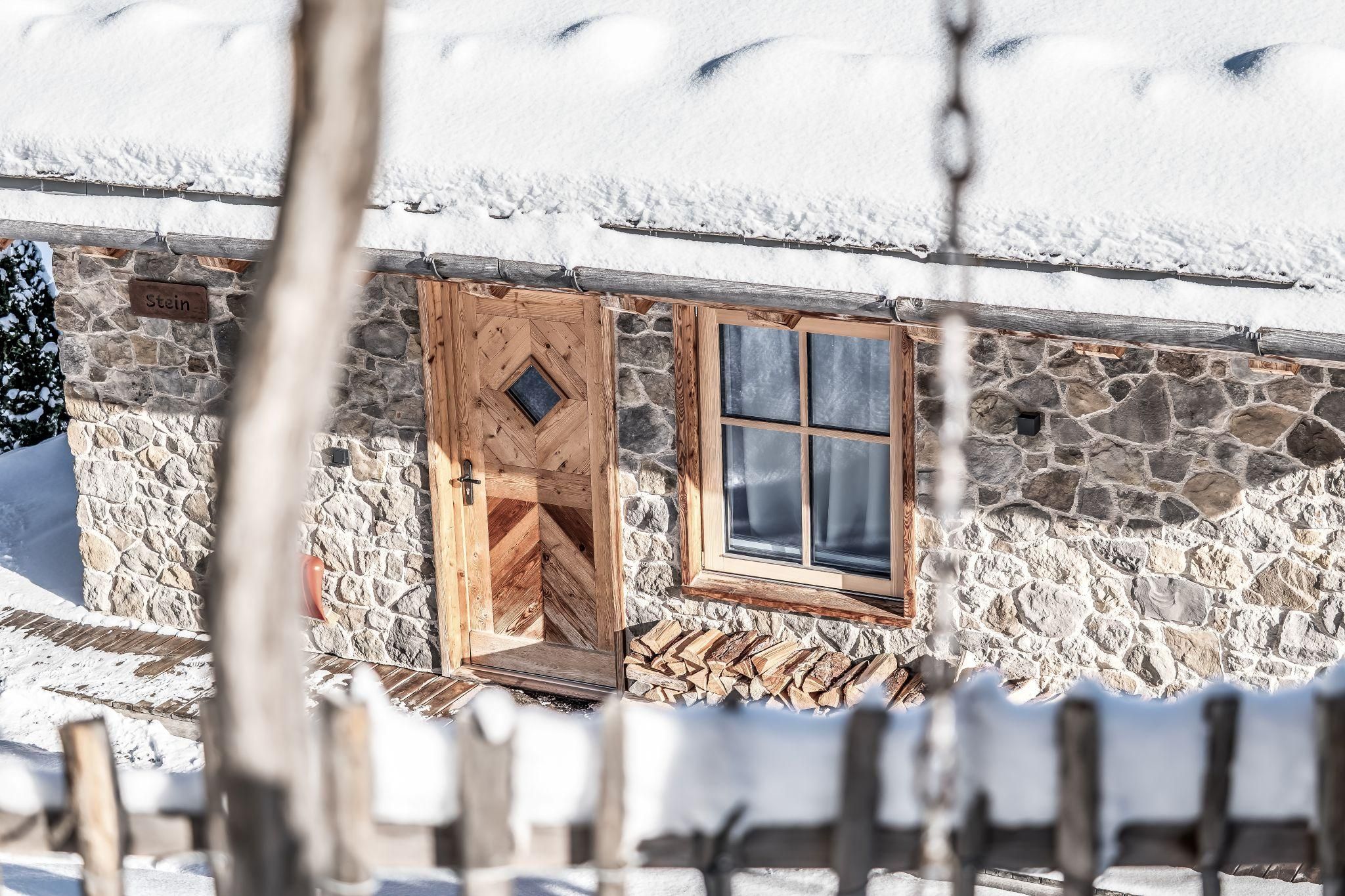 Eine gemütliche Stein Chalet im Schnee mit einem Holzofen und einer sichtbaren Eingangstür. Die Umgebung ist winterlich und ruhig.