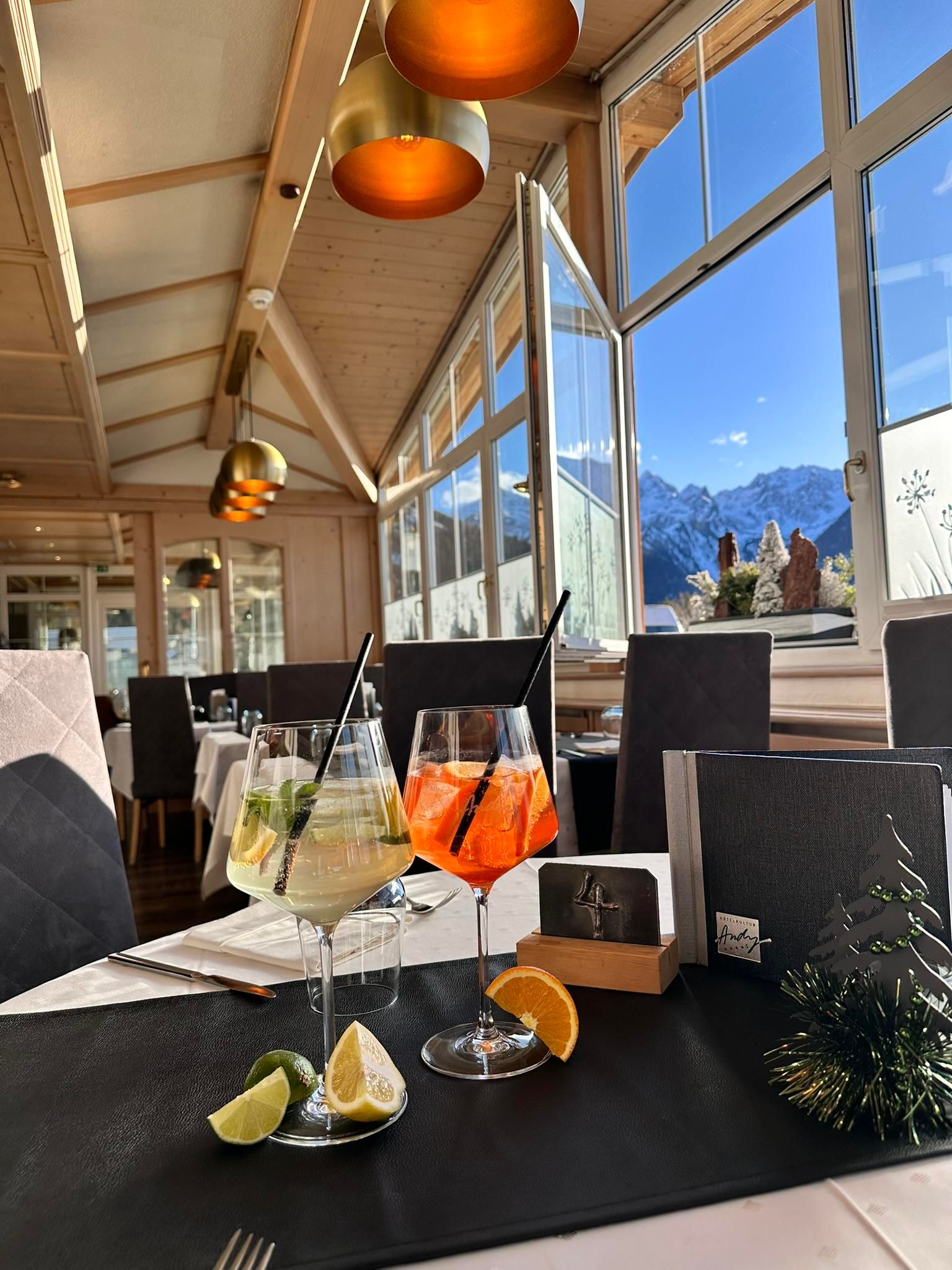 Ein stilvolles Restaurant mit einer Aussicht auf die Berge. Zwei bunte Cocktails stehen auf einem Tisch, umgeben von frischen Früchten.