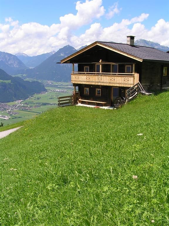 Ein gemütliches Holzhaus auf einer grünen Wiese mit Blick auf die Berge. Der Himmel ist klar und es ist sonnig.