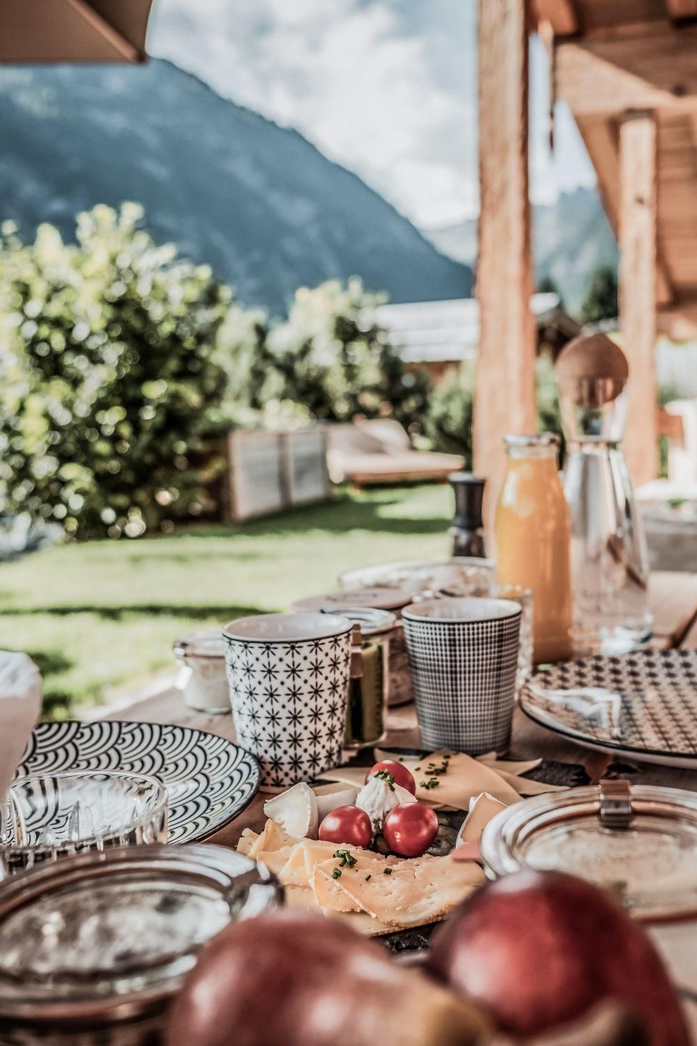 Ein schön gedeckter Tisch im Freien mit frischem Obst, Käse und Getränken. Im Hintergrund sind grüne Bäume und Berge zu sehen.