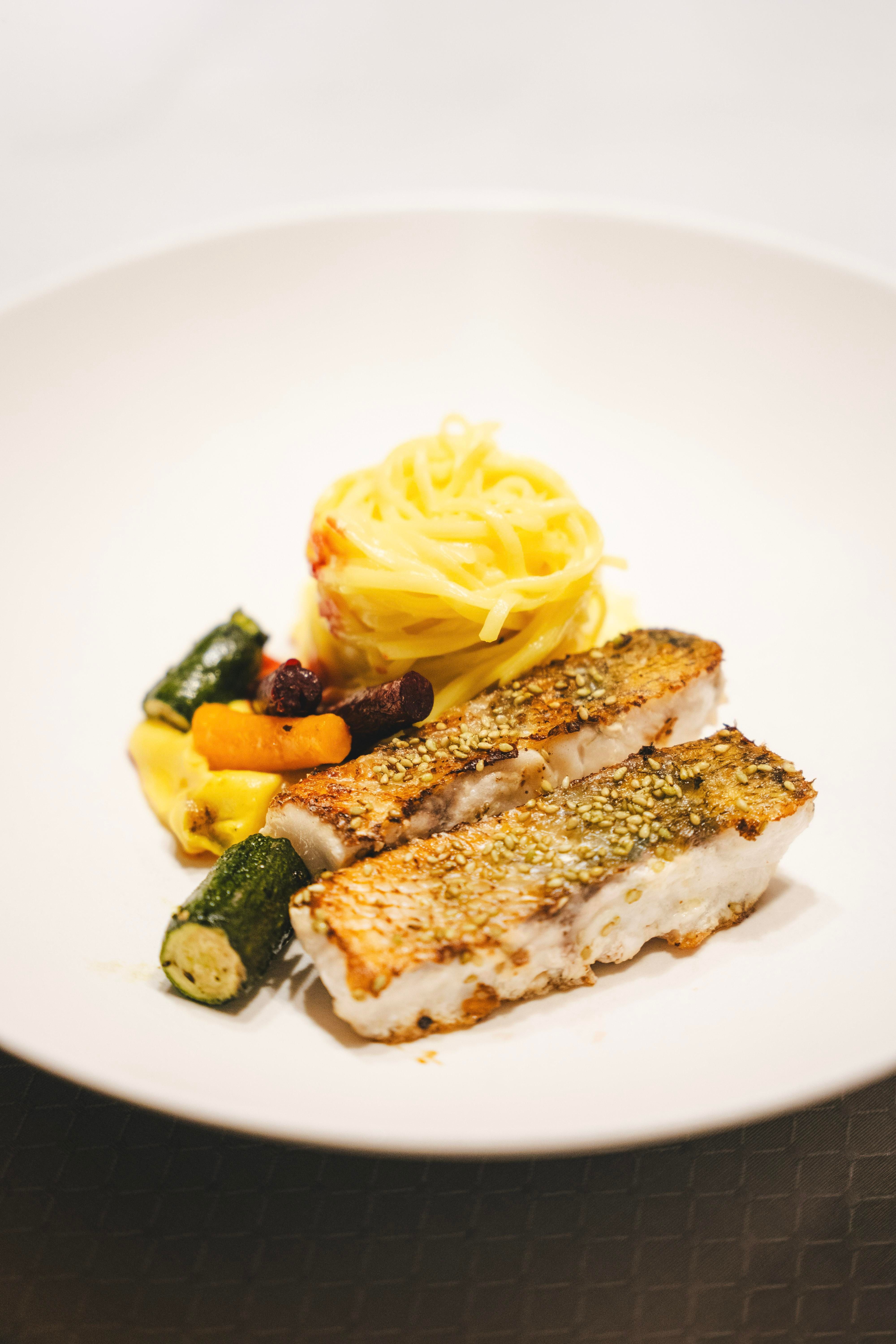 Ein Teller mit gebratenem Fisch, serviert mit Pasta und buntem Gemüse. Das Gericht sieht appetitlich und frisch aus.