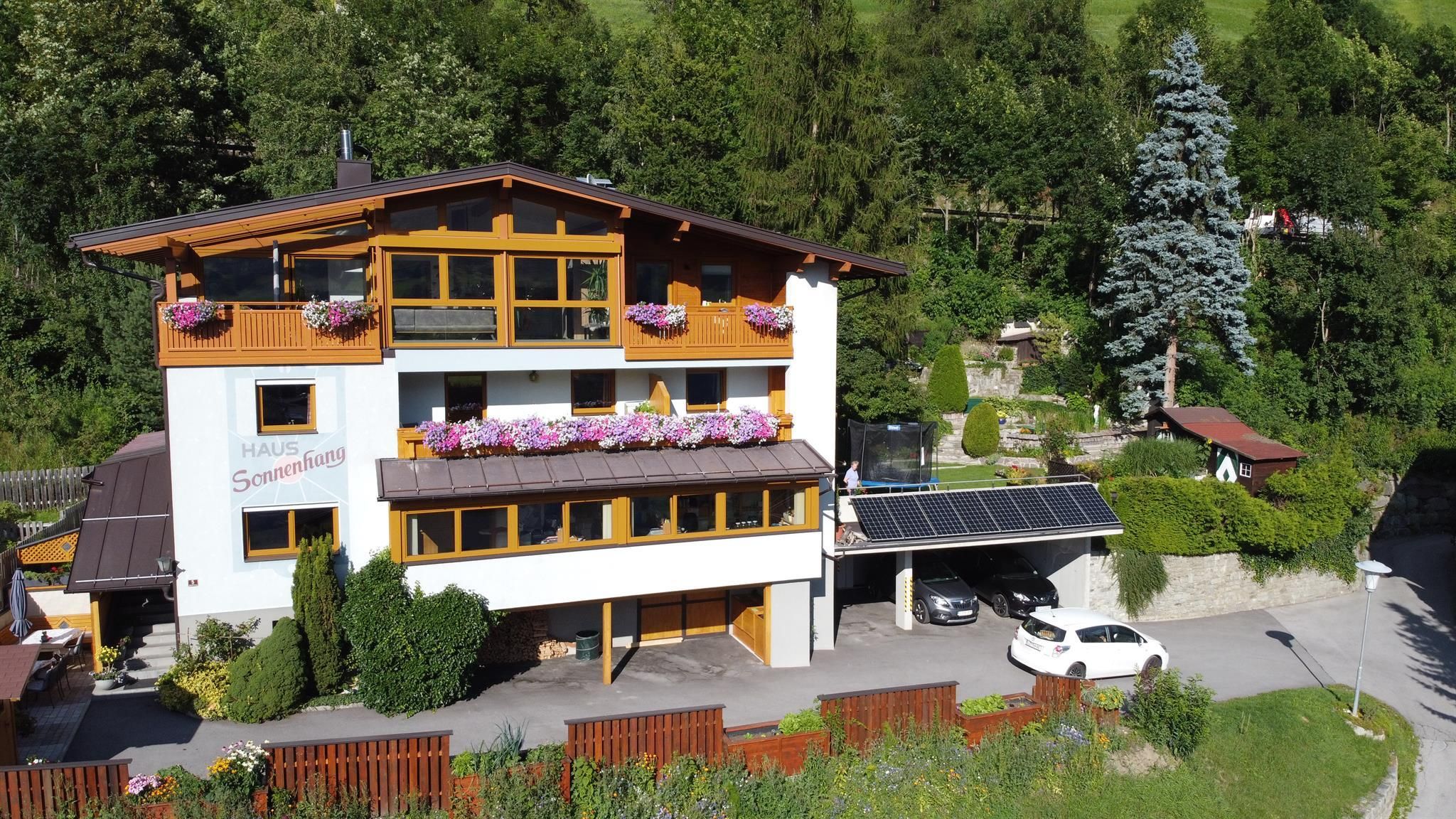 Ein mehrstöckiges Gästehaus im alpinen Stil mit Holzbalkonen und Blumenkästen, gelegen in grüner Hanglage mit angrenzendem Parkplatz und Carport.