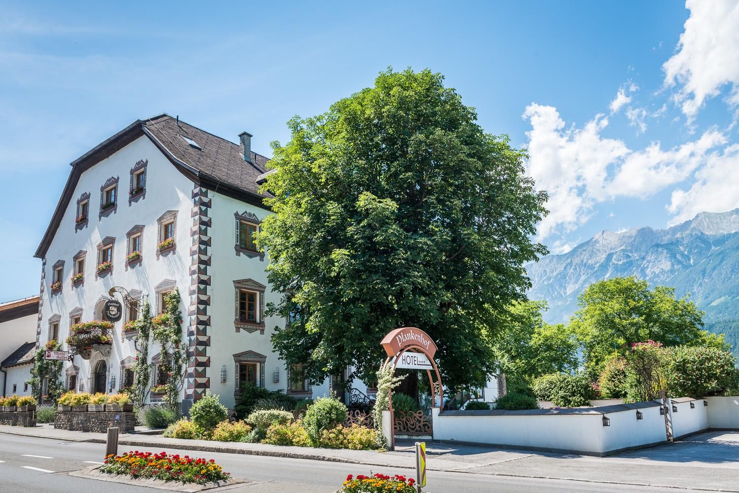 Ein charmantes Gebäude im traditionellen Stil, umgeben von Bäumen und Blumen. Im Hintergrund sind Berge und ein blauer Himmel zu sehen.