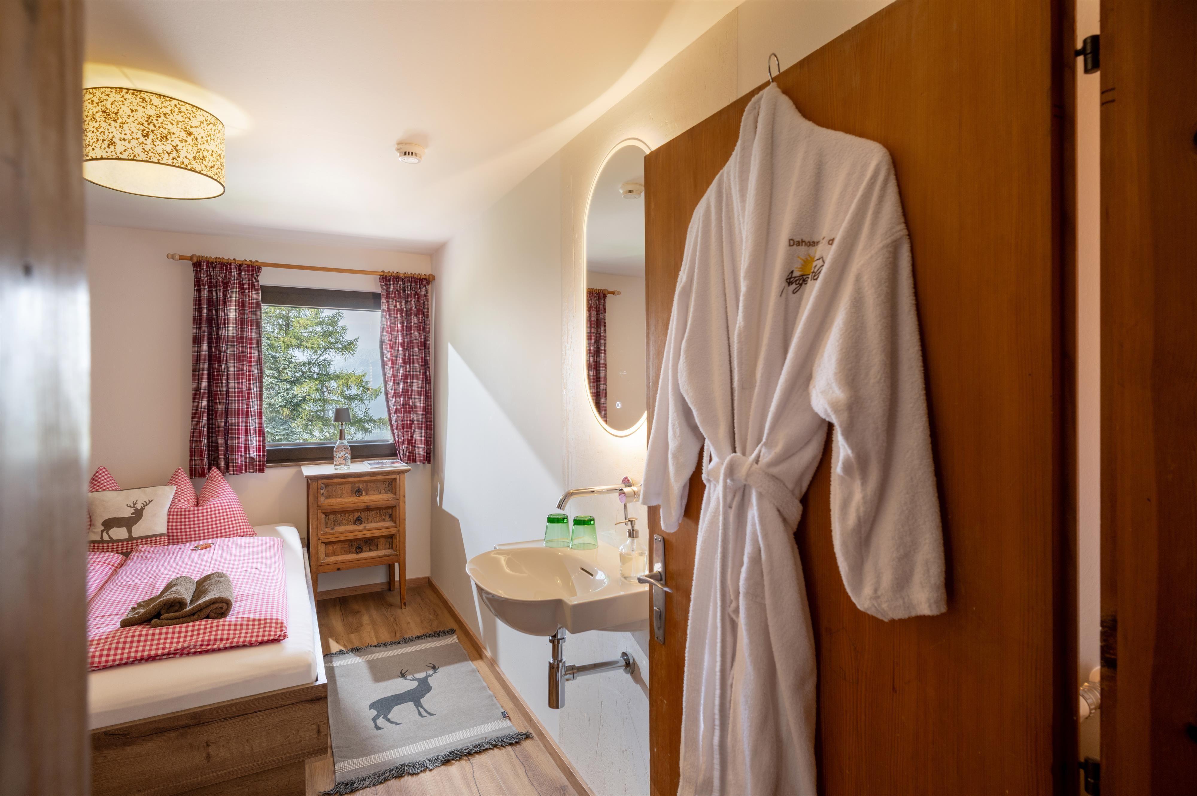 Ein gemütliches Gästezimmer mit einem Doppelbett und einem Fenster, das viel Licht hereinlässt. Ein Bademantel hängt an der Tür, und es gibt eine kleine Waschgelegenheit im Raum.