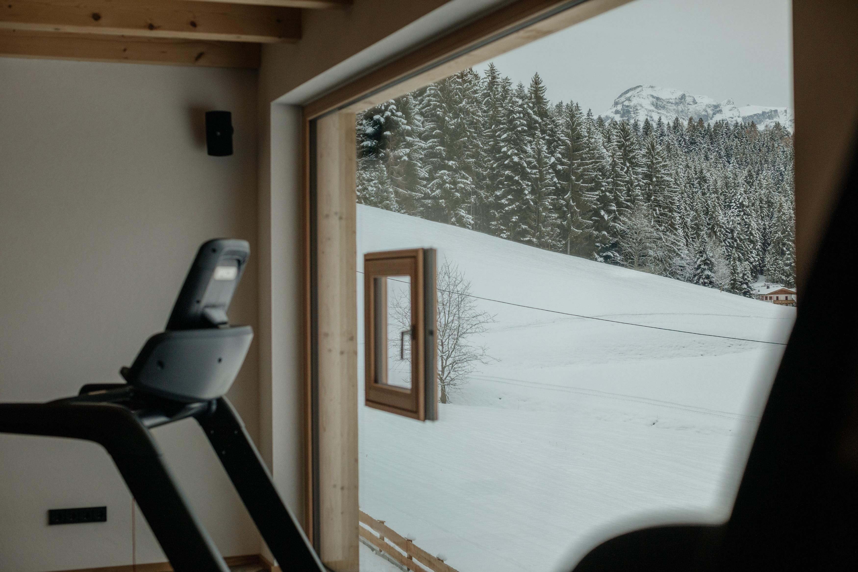 Ein Fitnessraum mit einem Blick auf eine schneebedeckte Landschaft und Nadelbäume. Durch das Fenster strahlt der Winterzauber herein.