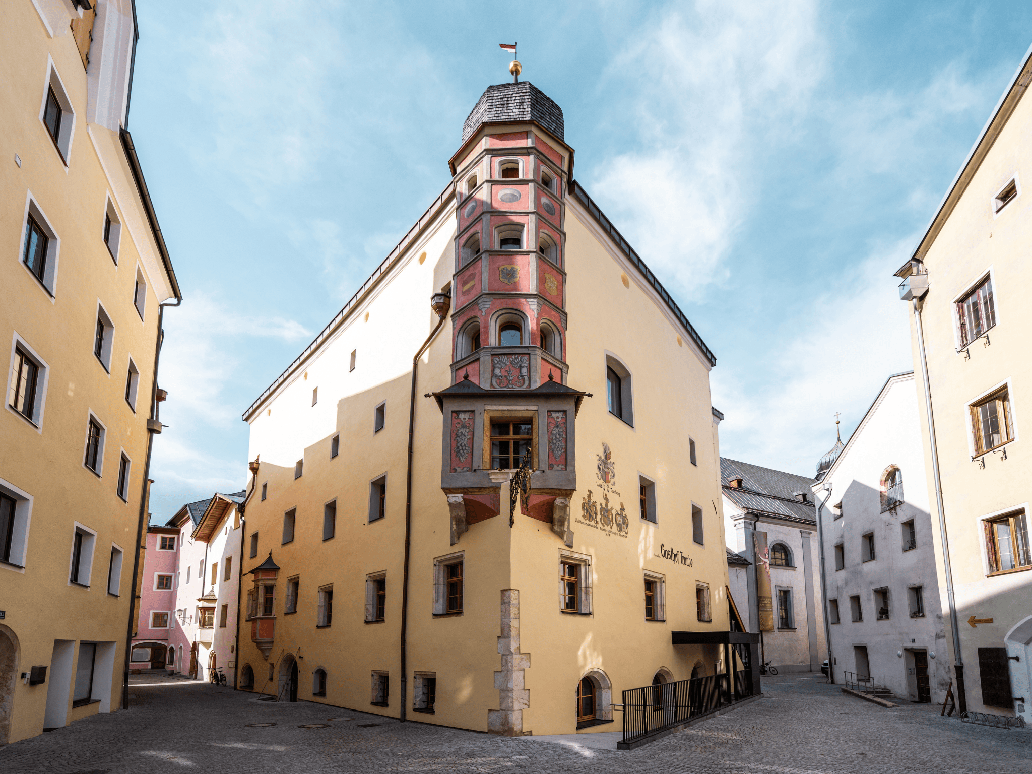 Ein historisches Gebäude mit bunten Fresken und einer markanten Fassade. Es steht zwischen schmalen, hellen Straßen in einer charmanten Stadt.