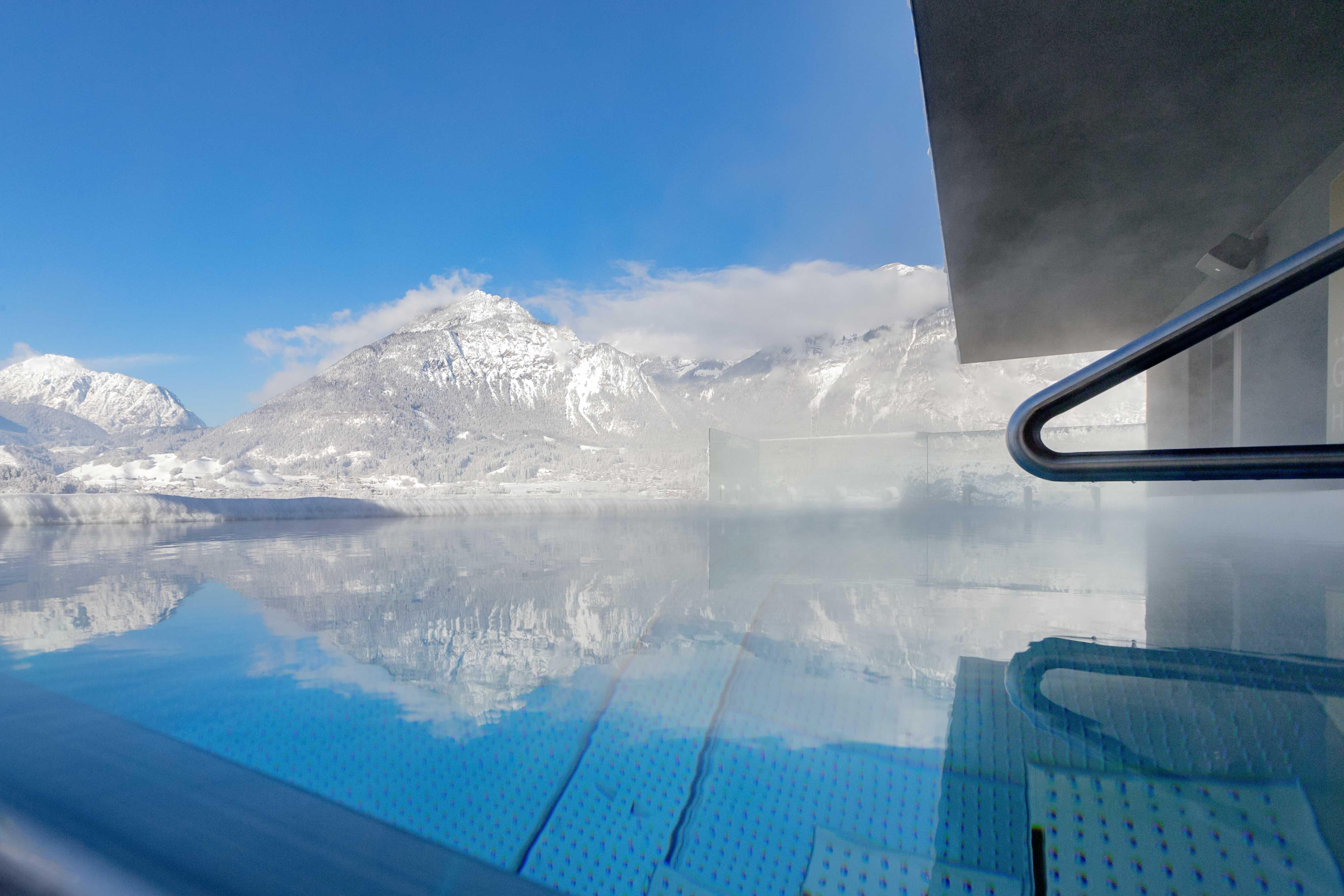 Ein Infinity-Pool mit Blick auf schneebedeckte Berge unter einem klaren blauen Himmel. Die ruhige Wasseroberfläche spiegelt die umliegende Berglandschaft wider.