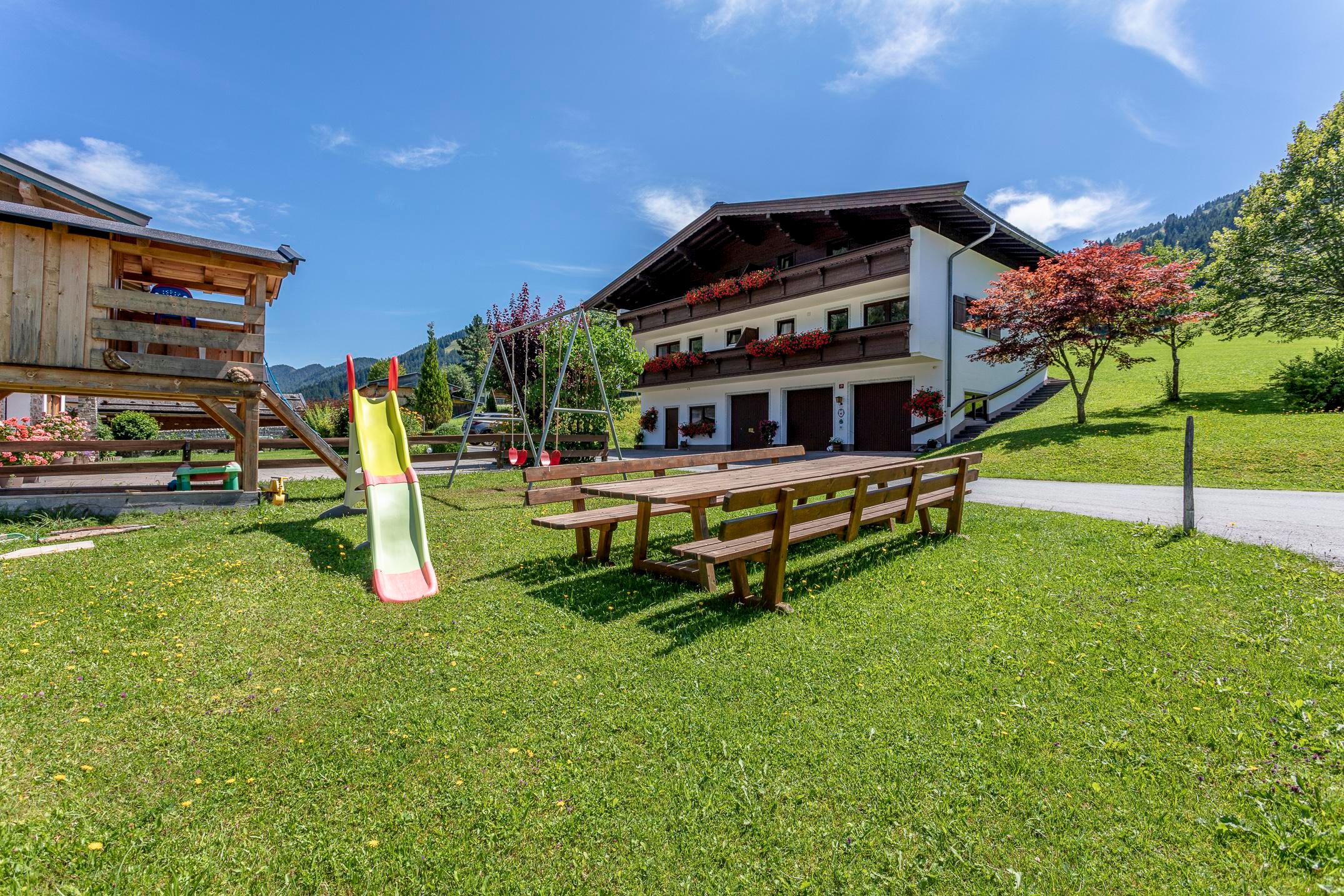 Ein gepflegter Garten mit einer Holzrutsche und Sitzbänken. Im Hintergrund steht ein gemütliches Gebäude umgeben von Bäumen.