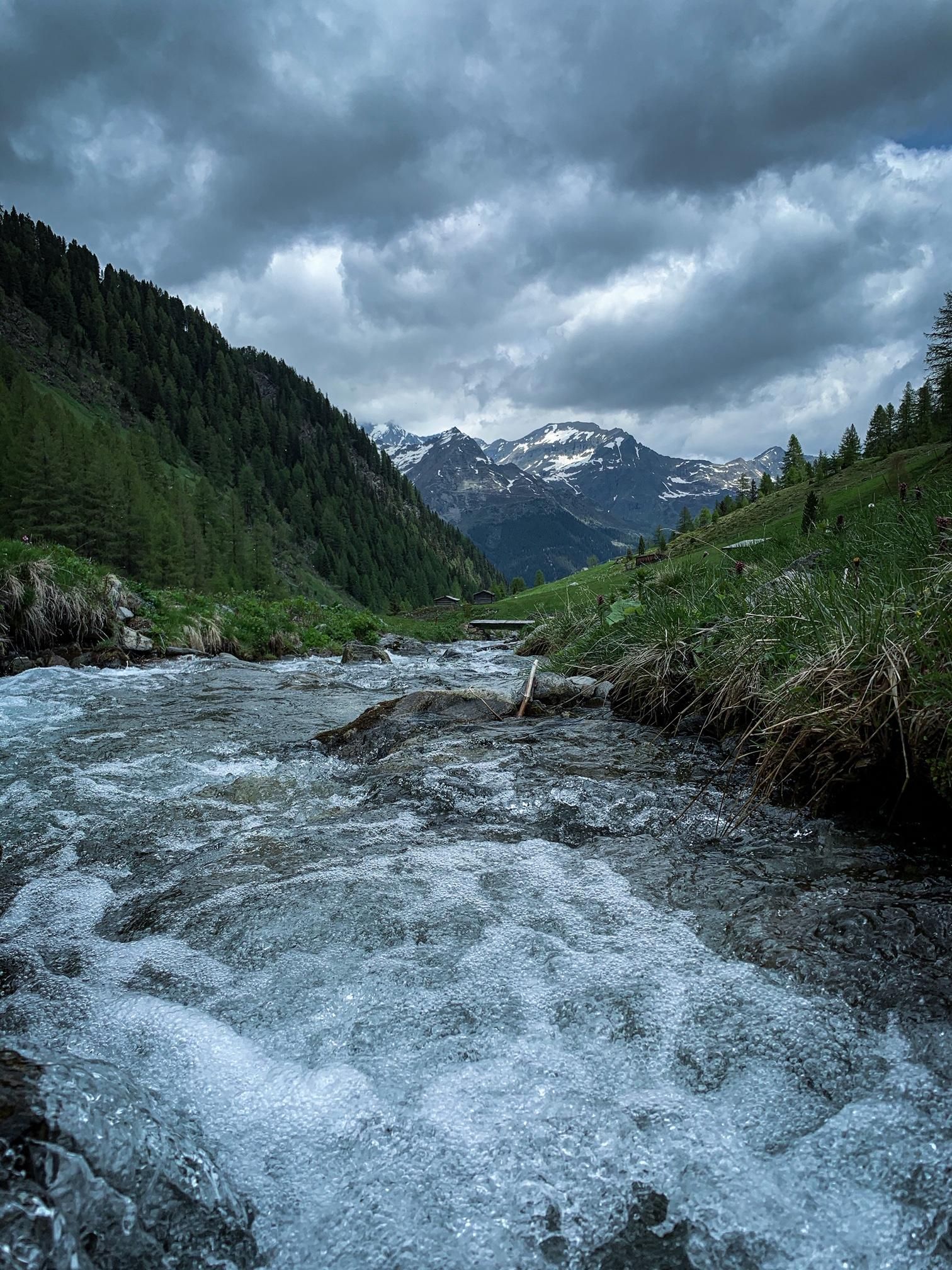 Ein klarer Fluss fließt durch eine grüne Berglandschaft. Im Hintergrund sind schneebedeckte Berge und bewölkter Himmel zu sehen.