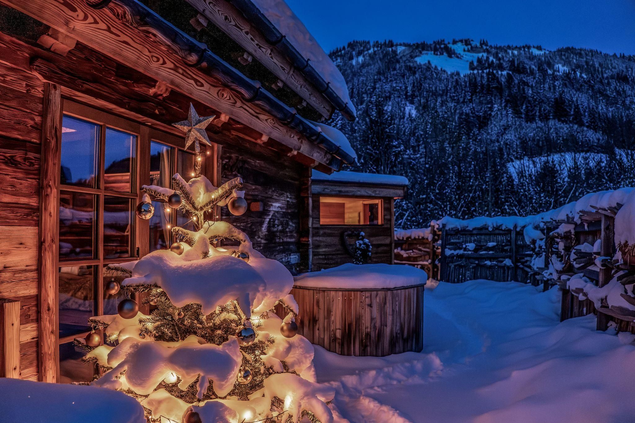 Eine winterliche Landschaft mit einem Weihnachtsbaum, der mit Lichtern geschmückt ist. Im Hintergrund sind schneebedeckte Berge und ein Chalet zu sehen.