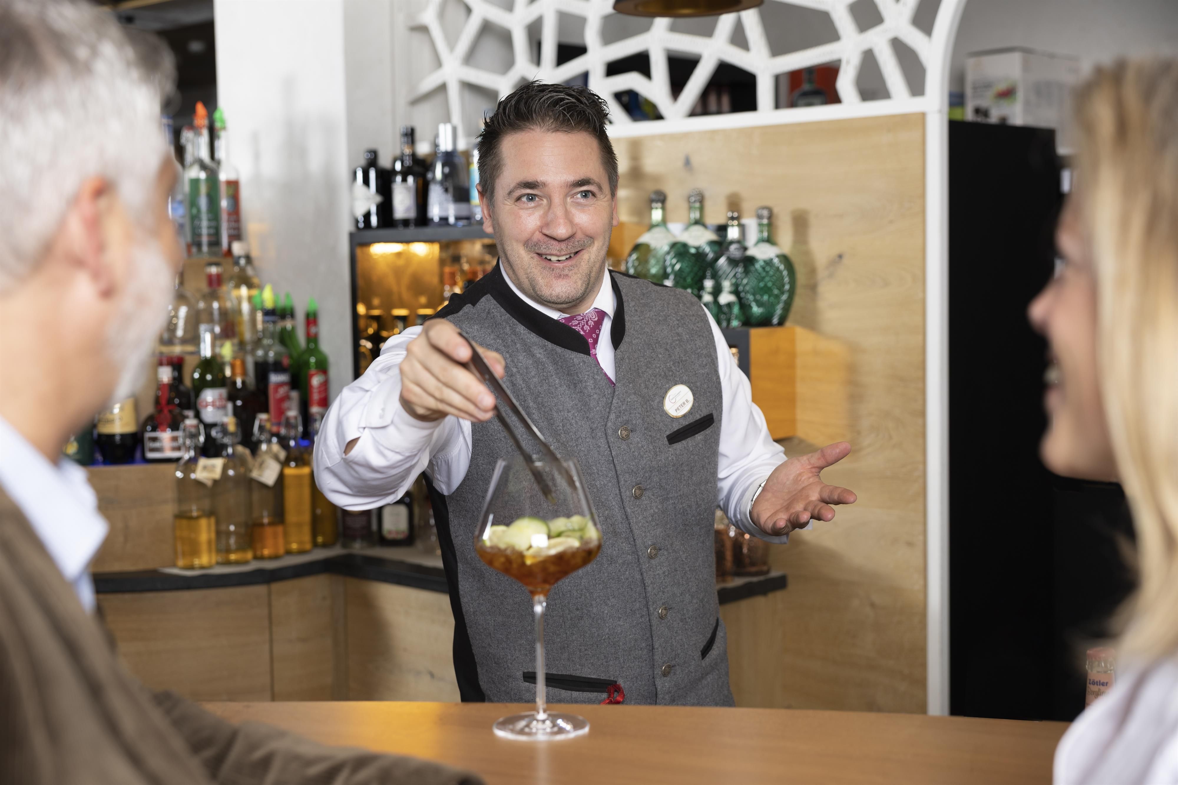Ein Barkeeper serviert einen Cocktail an Gäste. Der Barkeeper lächelt freundlich und präsentiert das Getränk mit einer einladenden Geste.