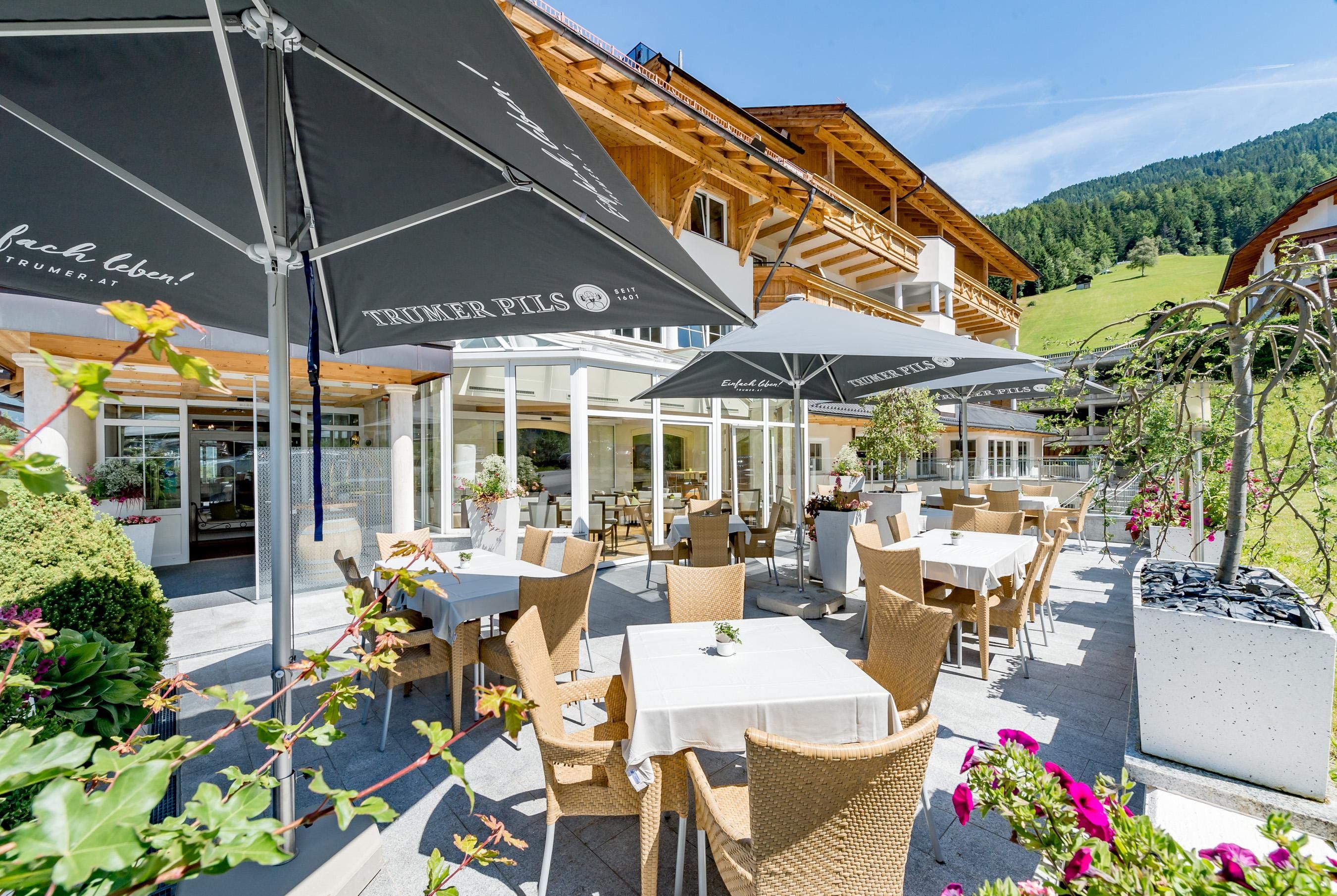 Eine einladende Terrasse mit Tischen und Stühlen, umgeben von Blumen. Im Hintergrund ist ein traditionelles Gebäude in malerischer Landschaft zu sehen.
