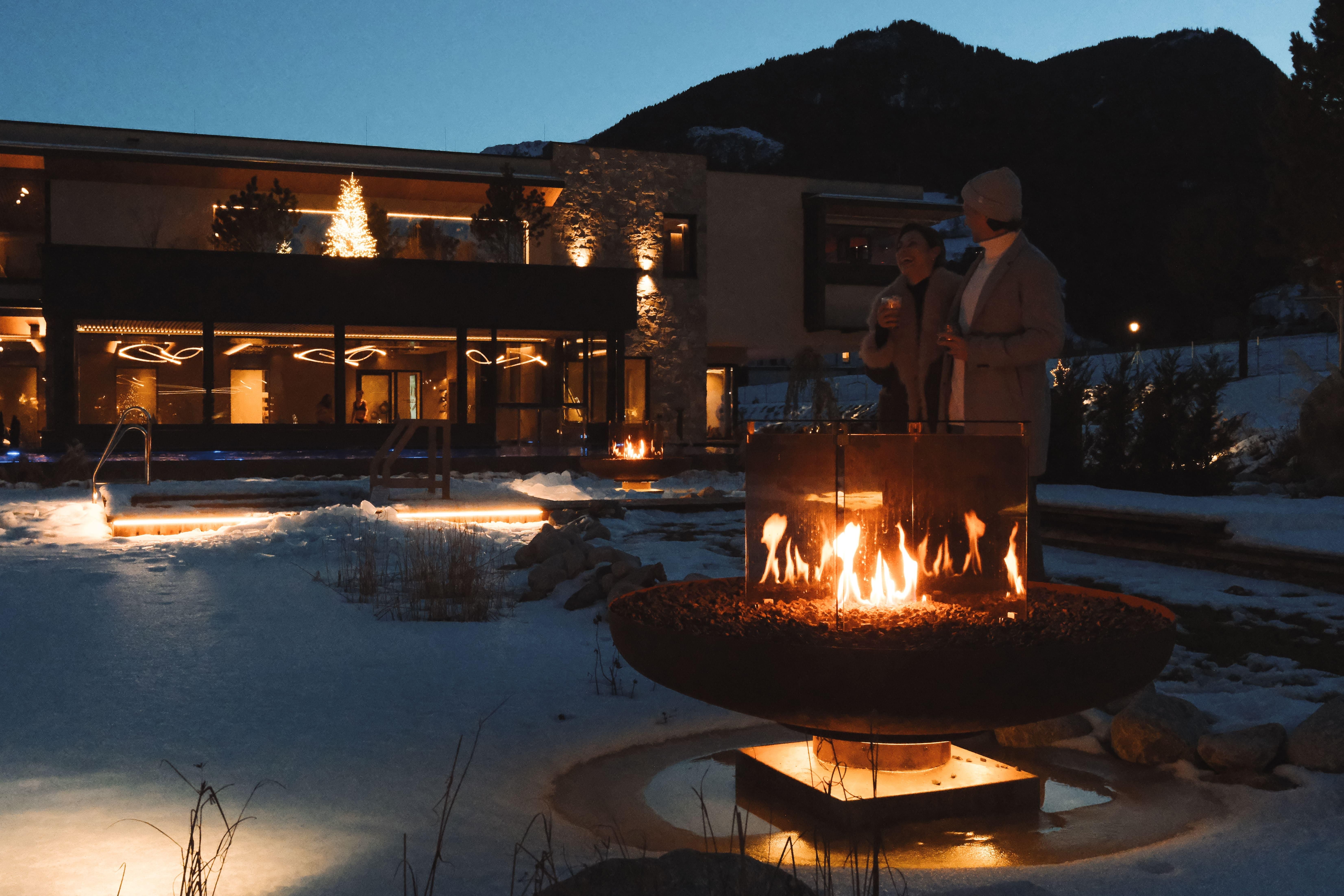 Eine gemütliche Winterlandschaft mit einem Feuer in einem großen Metallring. Im Hintergrund ist ein stilvolles Gebäude mit Weihnachtsbeleuchtung und schneebedeckter Umgebung zu sehen.