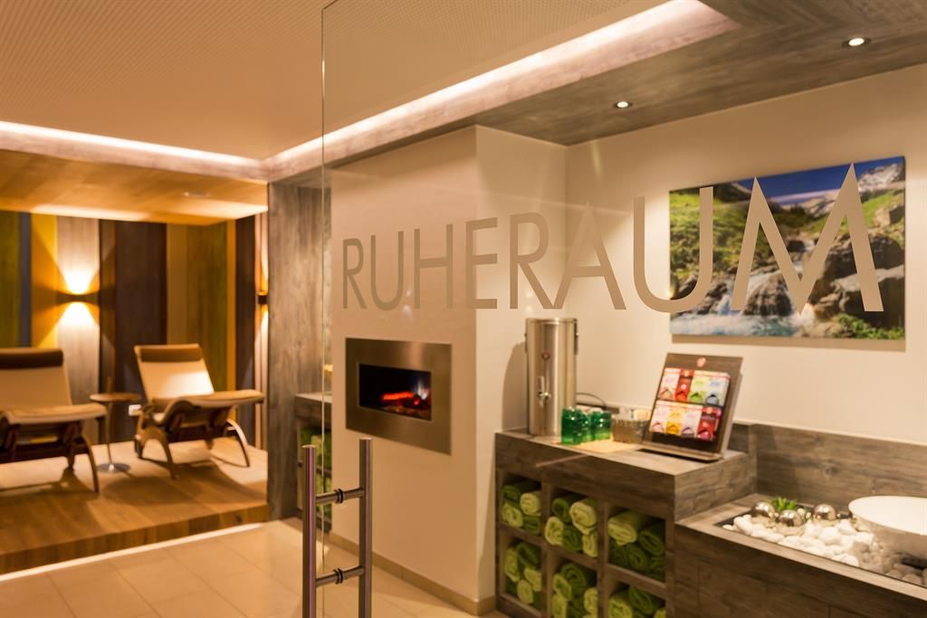 Ein ruhiger Raum mit einem modernen Design und gemütlichen Liegen. An der Wand befindet sich eine große Glasfläche mit der Aufschrift "RUHERAUM" und einem schönen Bild von Wasserfällen.