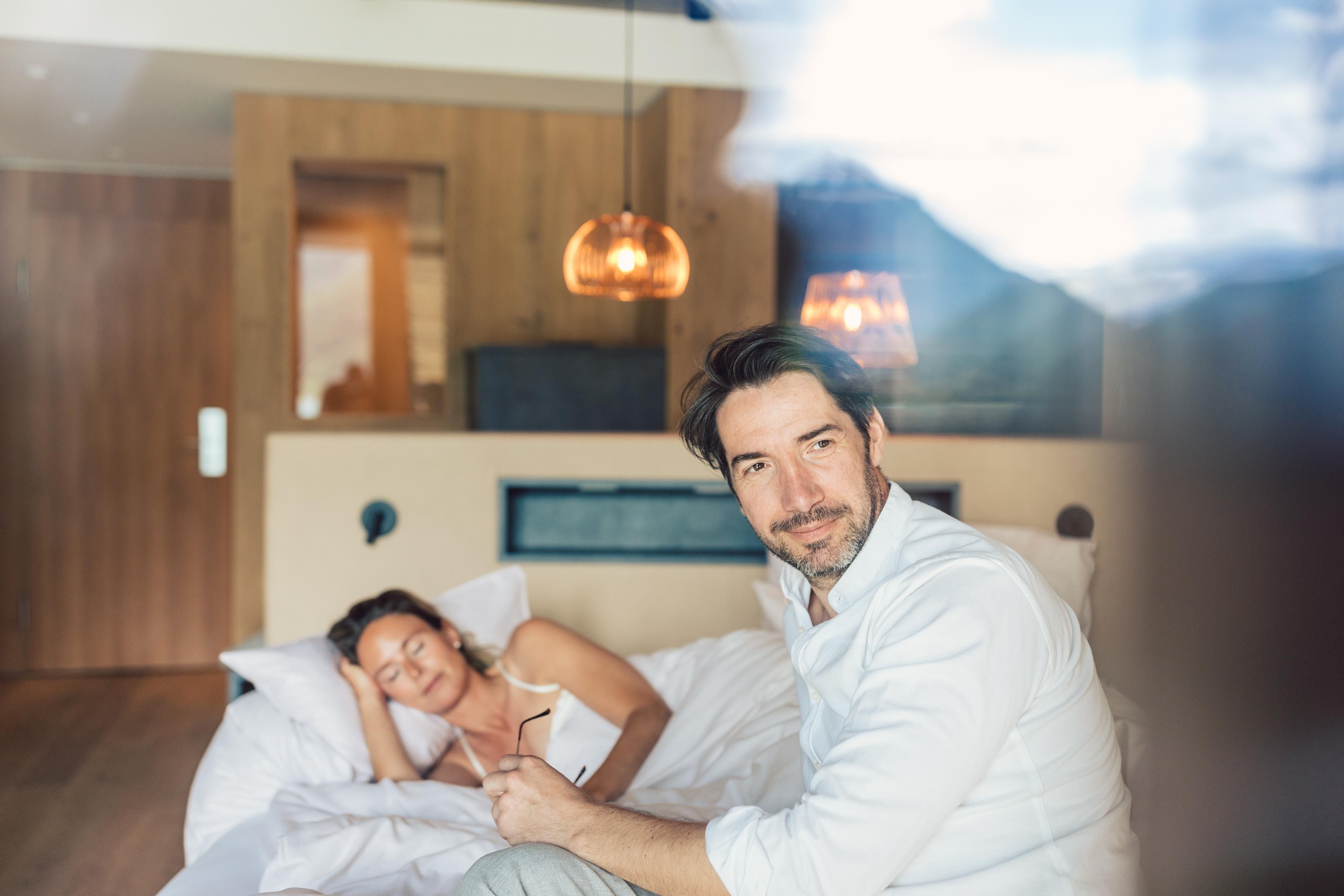 Ein Mann sitzt lächelnd auf dem Bett, während eine Frau daneben schläft. Der Raum hat eine moderne, gemütliche Atmosphäre mit Holzdetails und warmem Licht.