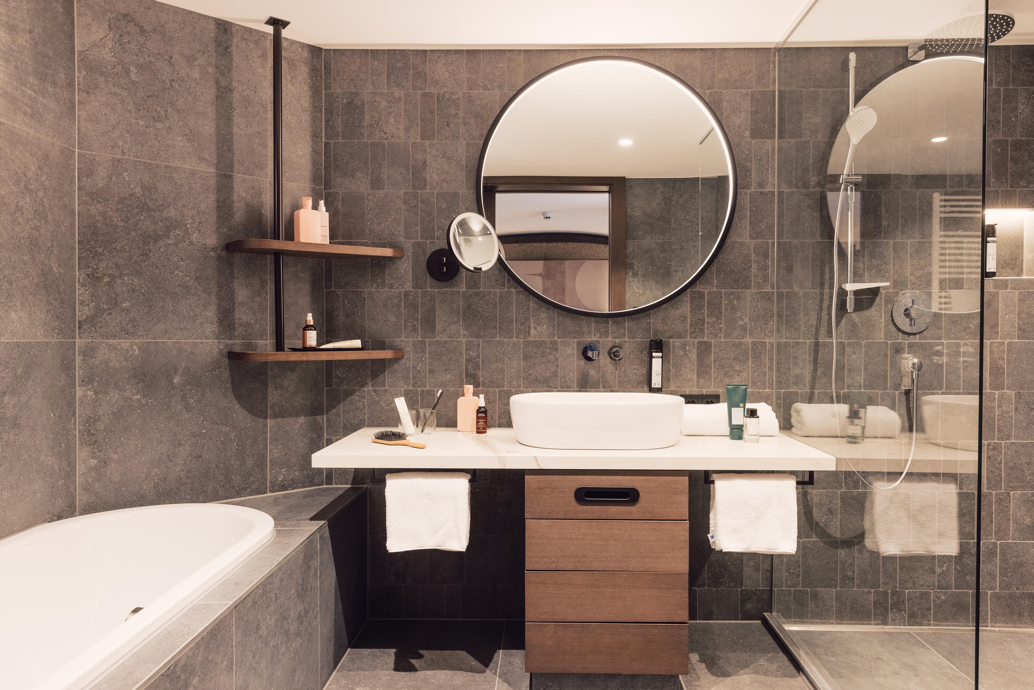 Ein modernes Badezimmer mit grauen Fliesen, einem runden Spiegel und einer Dusche. Die elegante Badewanne und die Regale sorgen für ein stilvolles Ambiente.