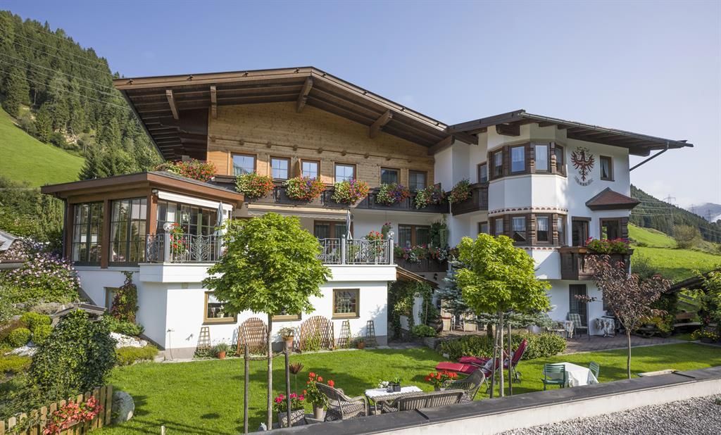 Ein charmantes alpenländisches Haus mit einem schönen Garten. Im Hintergrund sind grüne Berge und ein klarer Himmel zu sehen.