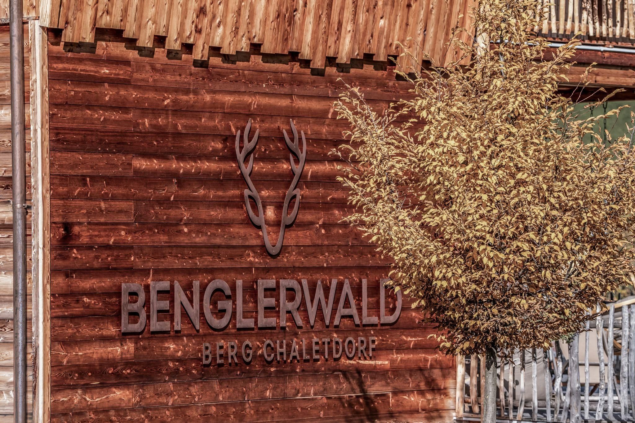 Ein Holzschild mit dem Schriftzug "Benglerwald Berg-Chaletdorf" und einem Geweih-Symbol. Im Vordergrund steht ein Baum mit gelben Blättern.