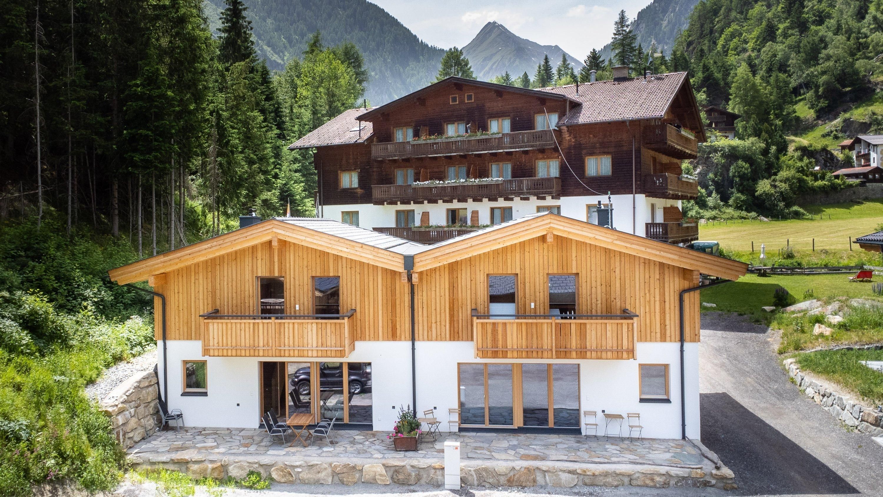 Zwei moderne Holzhäuser umgeben von grünen Bäumen und Bergen. Im Hintergrund ist ein traditionelles Gebäude zu sehen.