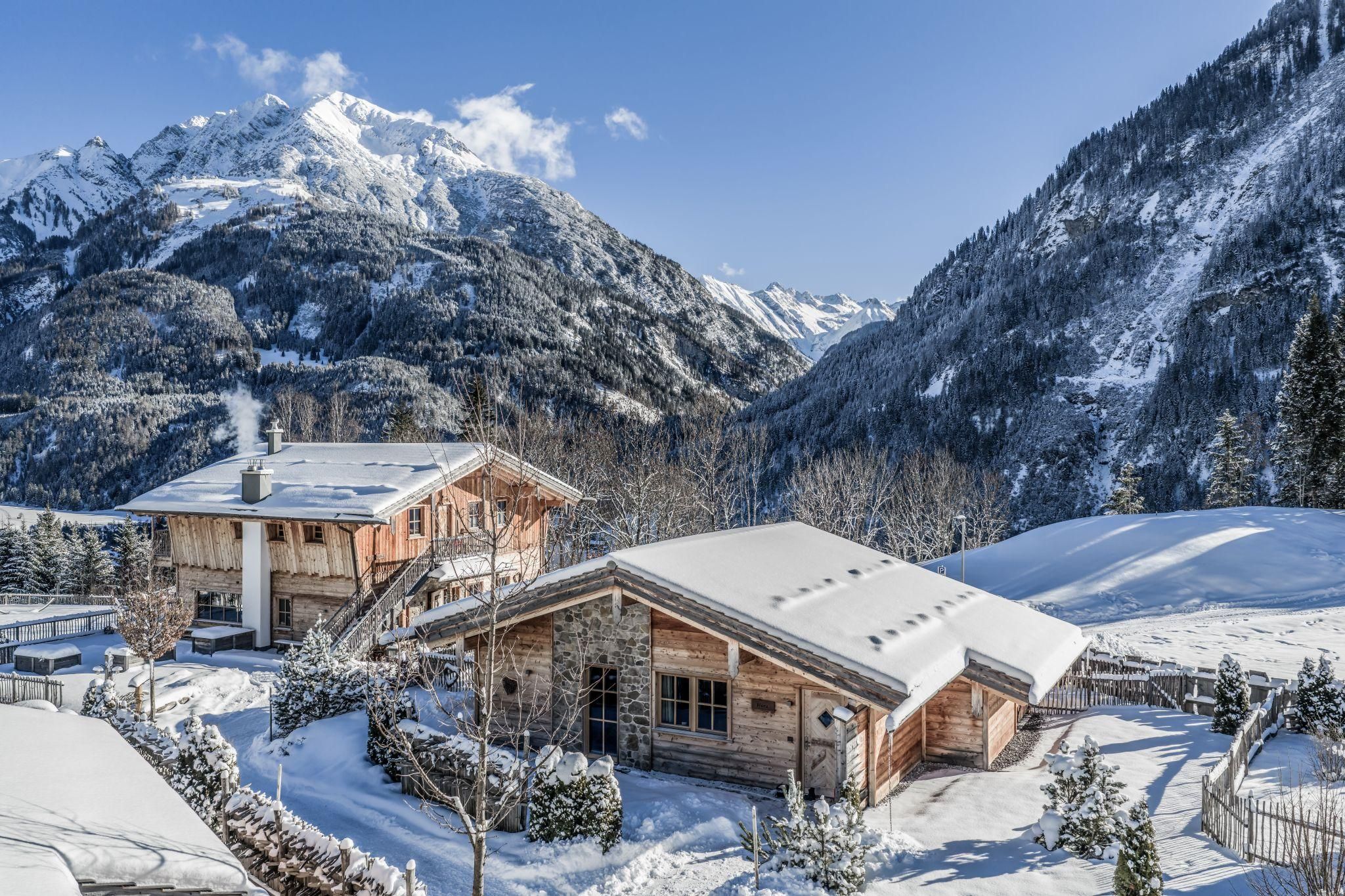 Eine Winterlandschaft mit verschneiten Chalets und majestätischen Bergen im Hintergrund. Der Himmel ist blau und die Sonne scheint auf die schneebedeckten Dächer.