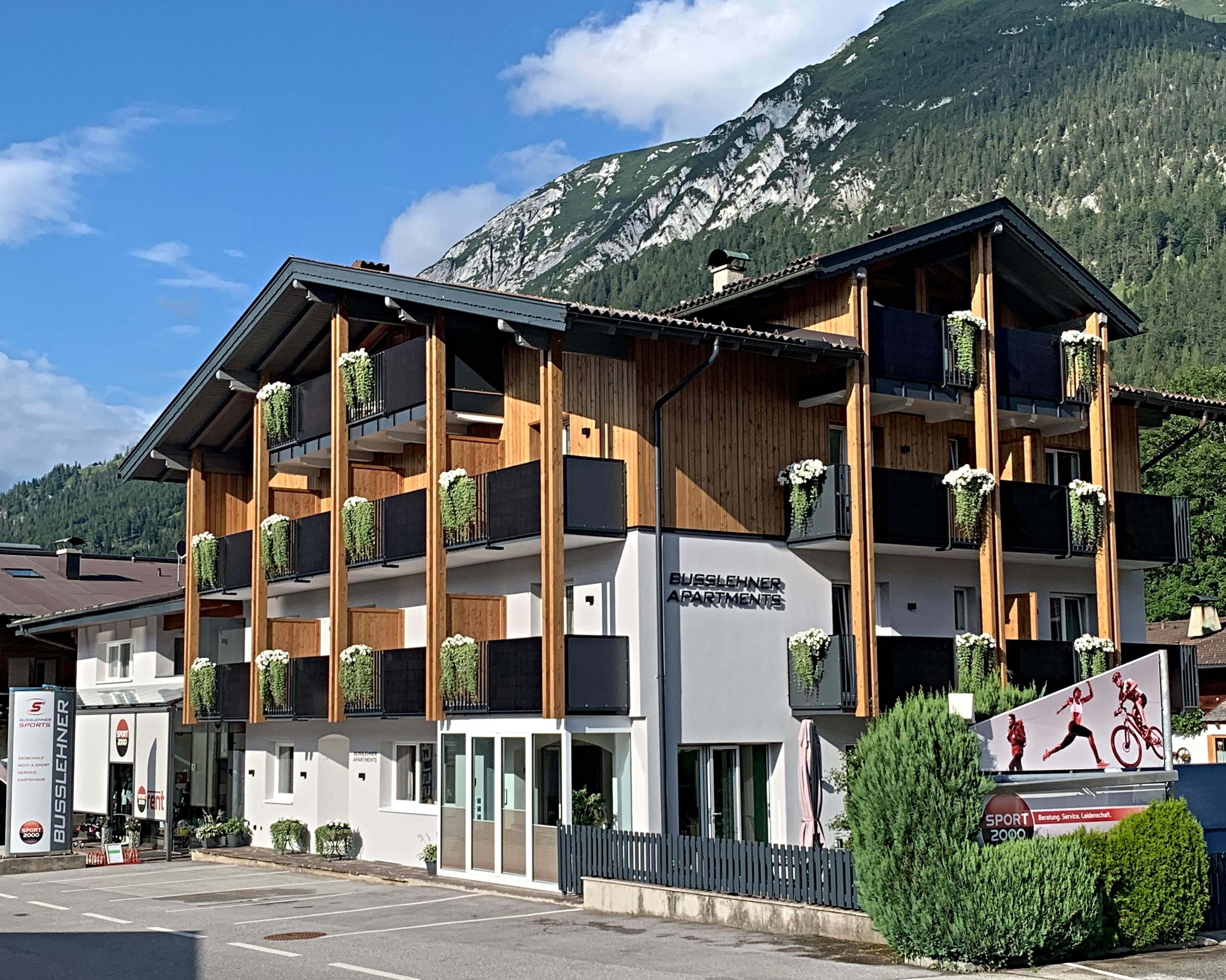 Ein modernes Gebäude im alpinen Stil mit Holzverkleidung und Balkonblumen. Im Hintergrund sind grüne Berge sichtbar.