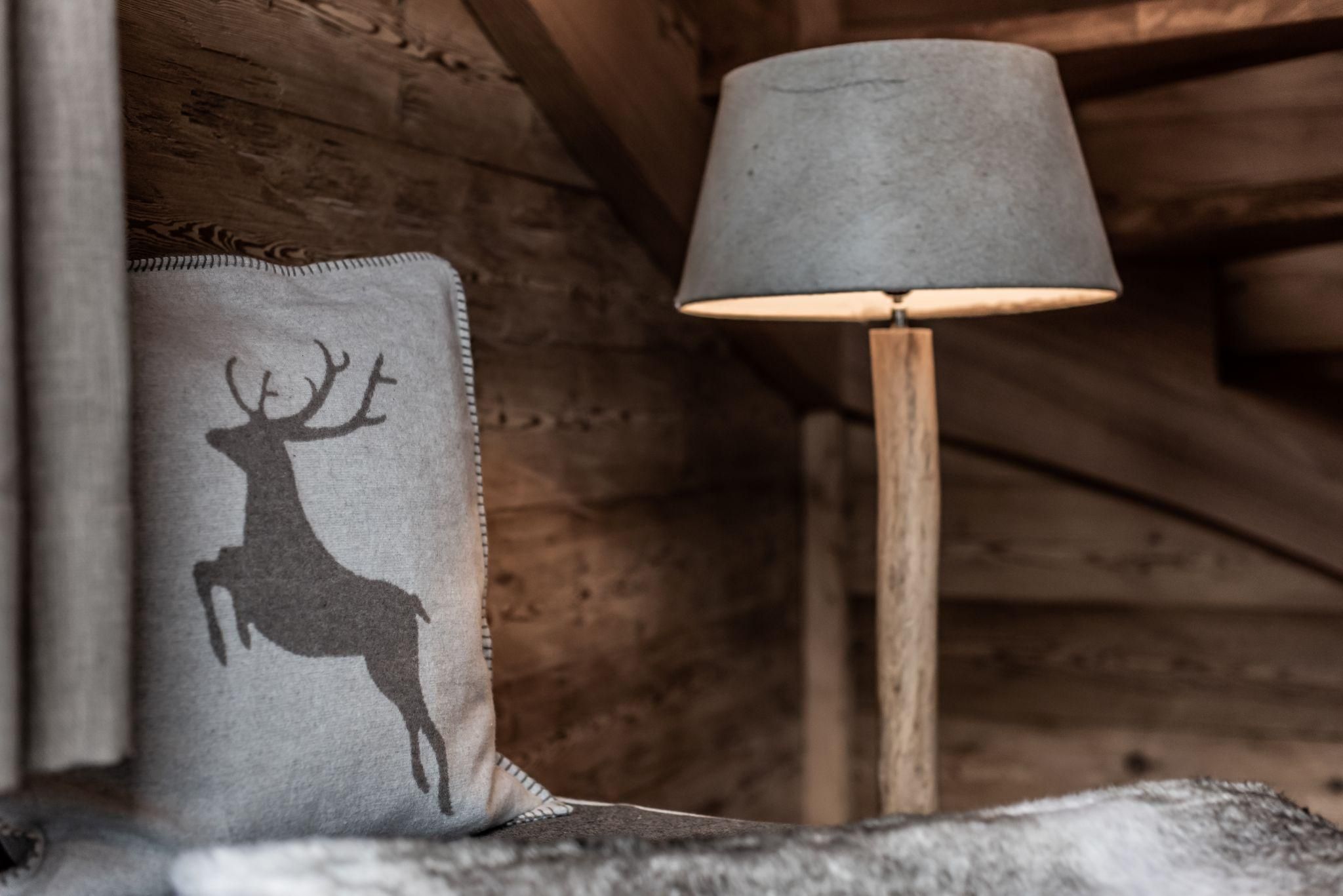 Ein behaglicher Raum mit einem Kissen, das ein Hirschmotiv zeigt, und einer Lampe aus Holz. Die Wände sind aus Holz und verleihen eine rustikale Atmosphäre.