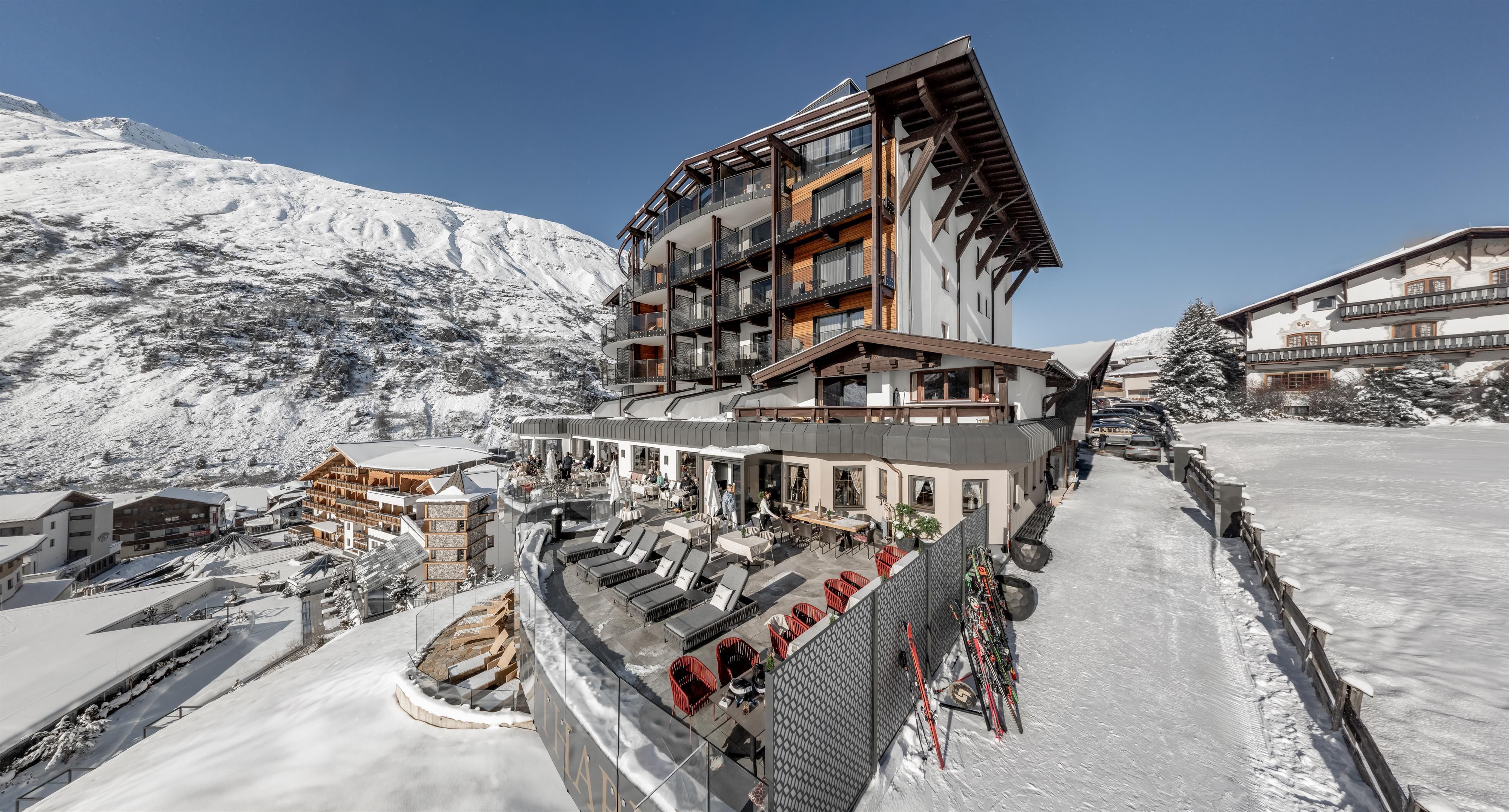 Ein modernes Hotel in einer verschneiten Berglandschaft. Klare blaue Himmel und gemütliche Liegen auf der Terrasse laden zum Entspannen ein.