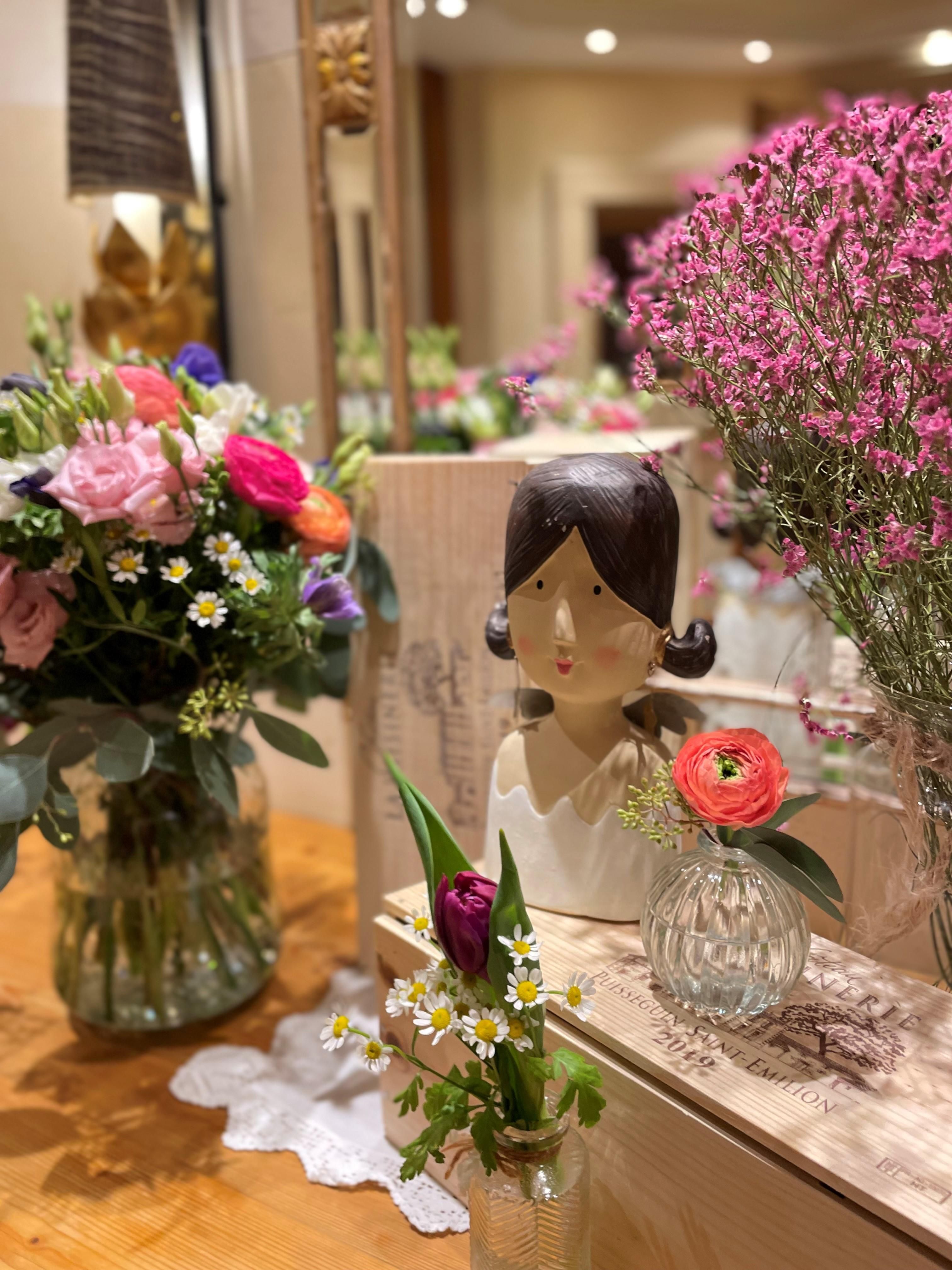 Ein hübsches Arrangement mit einem Blumengefäß und einer dekorativen Statue. Auf dem Tisch sind verschiedene Blumen und Pflanzen in klaren Vasen zu sehen.