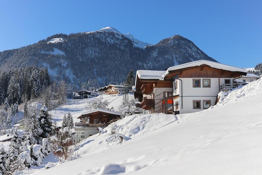 Eine malerische Winterlandschaft mit schneebedeckten Hügeln und gemütlichen Chalets. Im Hintergrund sind schneebedeckte Berge zu sehen.