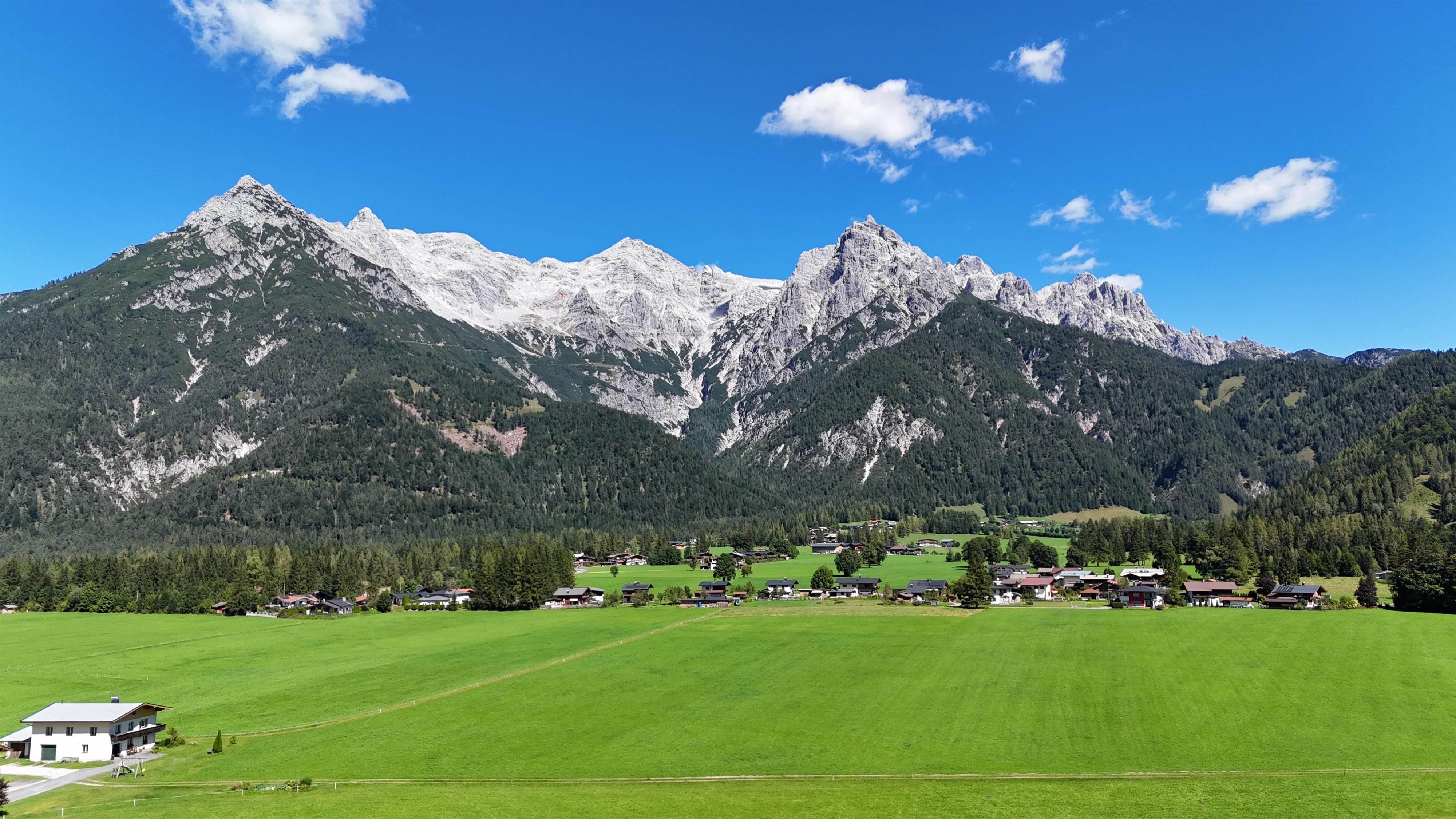 Eine beeindruckende Berglandschaft mit hohen Gipfeln und grünem Talboden. Im Vordergrund sind Wiesen und kleine Häuser sichtbar.