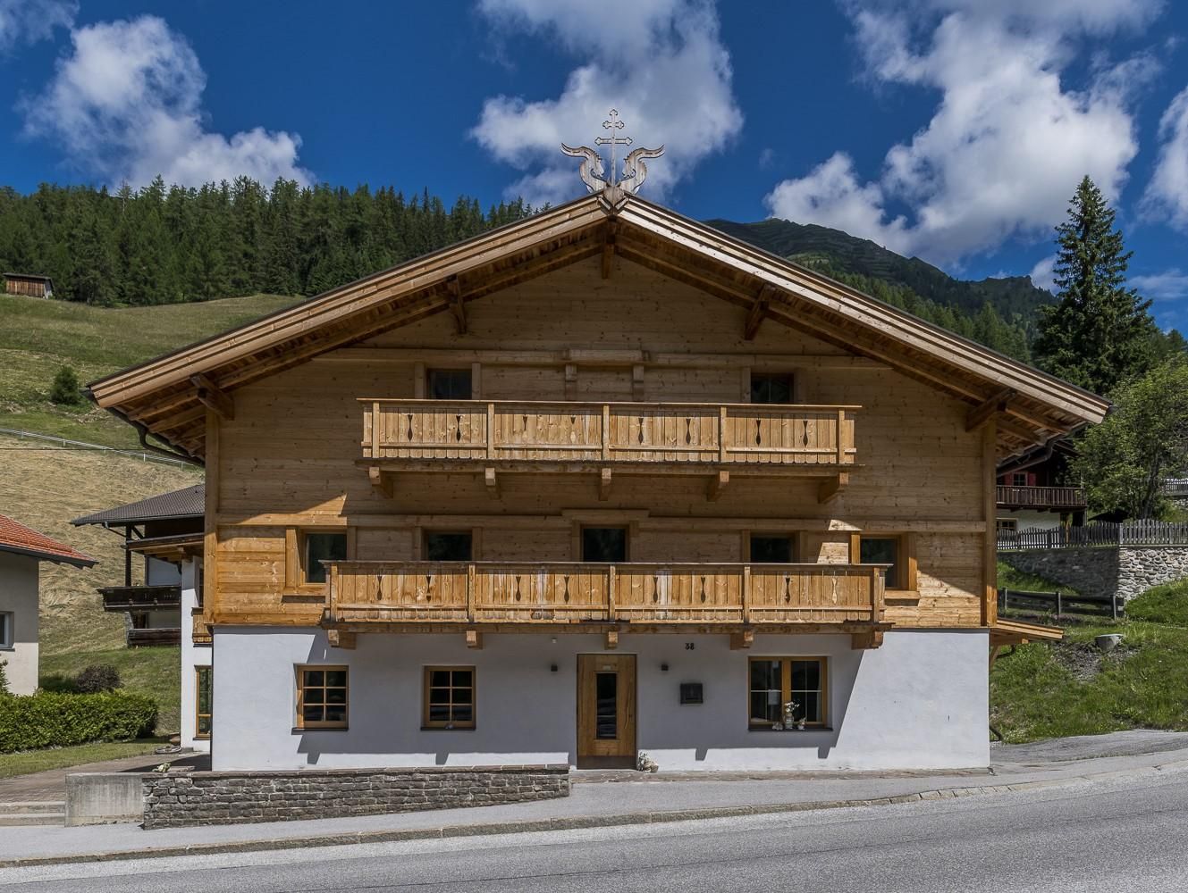 Ein traditionelles Holzhaus mit Balkon im Alpenstil. Im Hintergrund sind grüne Hügel und ein blauer Himmel mit einigen Wolken zu sehen.