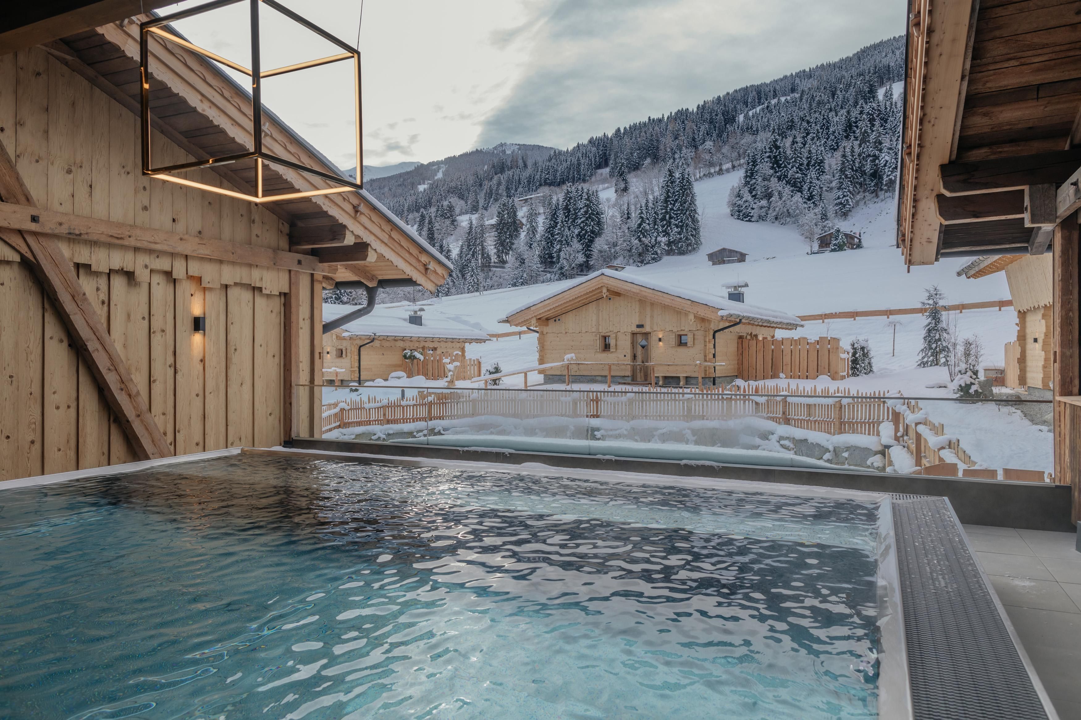Ein modernes Schwimmbad mit klarem Wasser, umgeben von einer winterlichen Landschaft. Im Hintergrund sind Holzchalets und schneebedeckte Berge zu sehen.