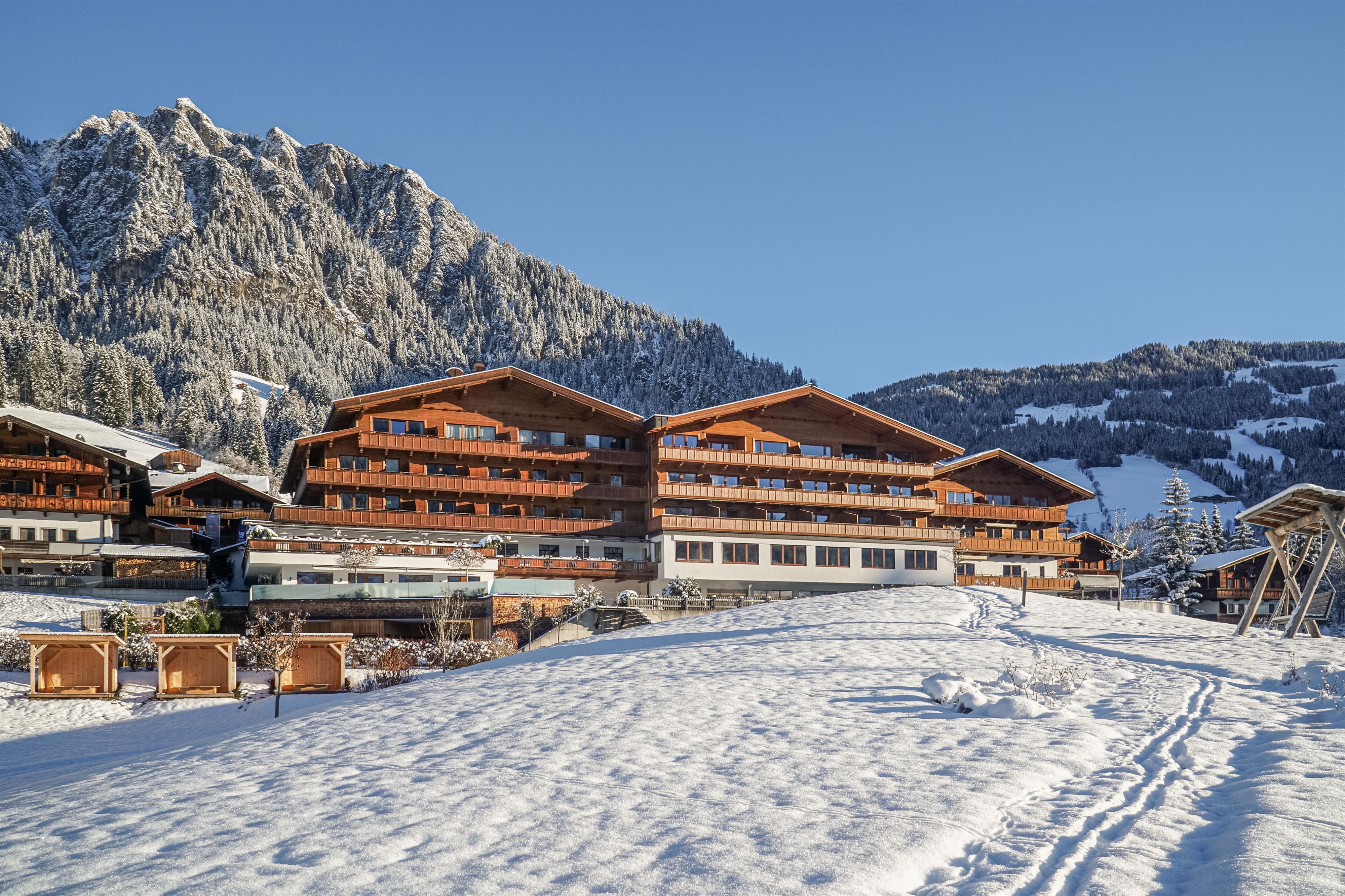 Ein traditionelles Holzgebäude in einer verschneiten Berglandschaft. Im Hintergrund sind hohe Berge und ein blauer Himmel zu sehen.