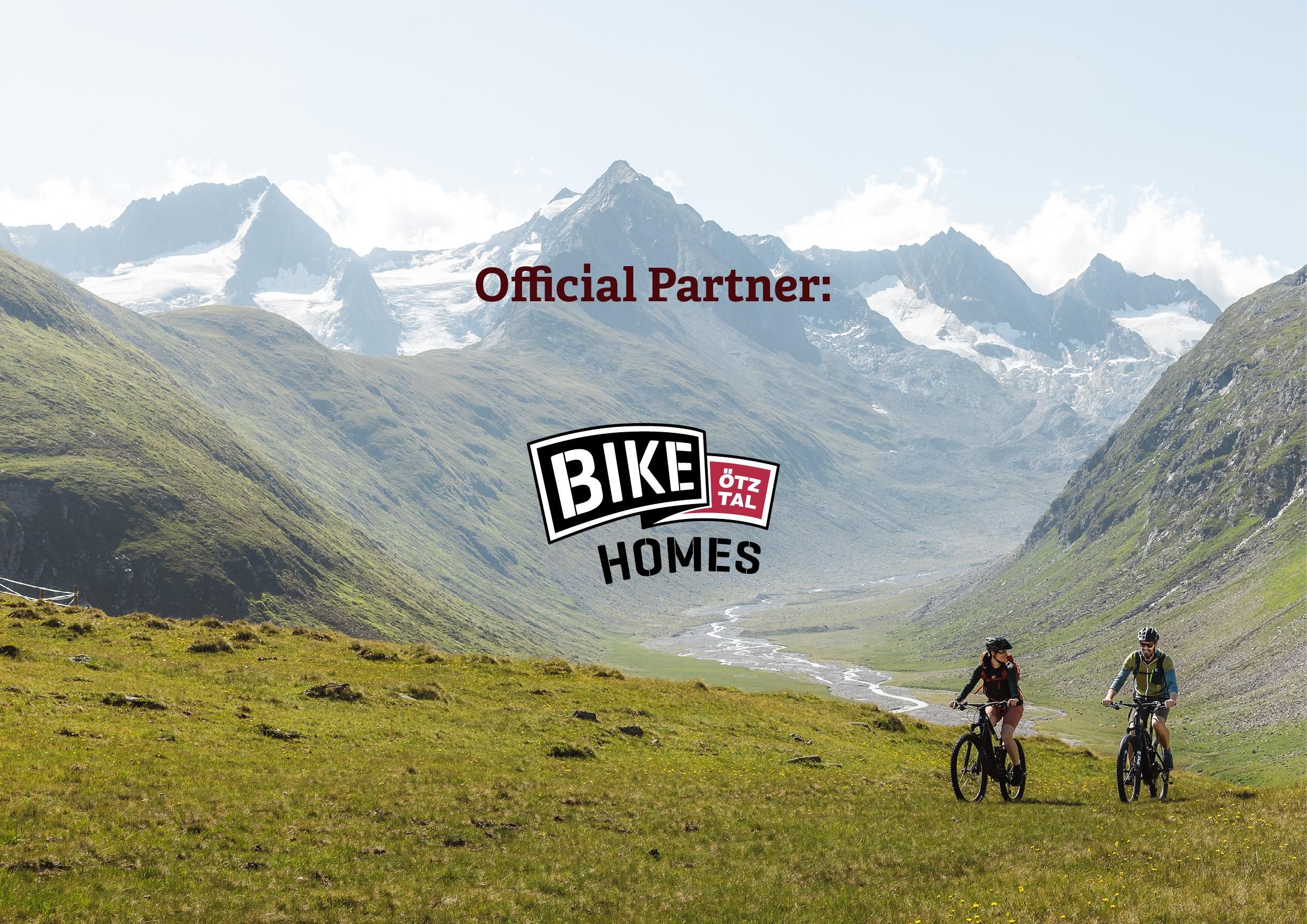 Zwei Radfahrer erkunden eine malerische Berglandschaft mit grünen Wiesen und beeindruckenden Bergen im Hintergrund. Das Logo „Bike Homes“ ist am unteren Bildrand zu sehen.