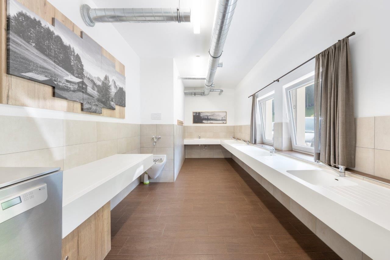 Ein modernes Badezimmer mit großzügigen Waschbecken und hellen Fliesen. Große Fenster sorgen für viel Tageslicht und eine angenehme Atmosphäre.