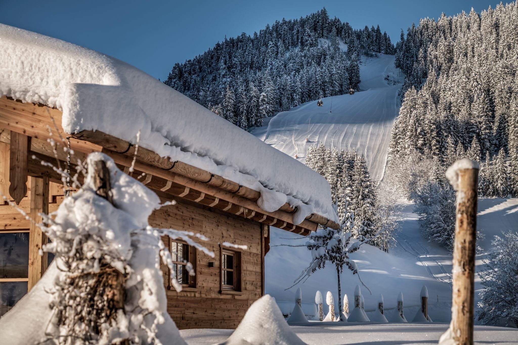 Eine gemütliches Chalet im Schnee mit einer dicken Schneeschicht auf dem Dach. Im Hintergrund sieht man verschneite Berge und Bäume.