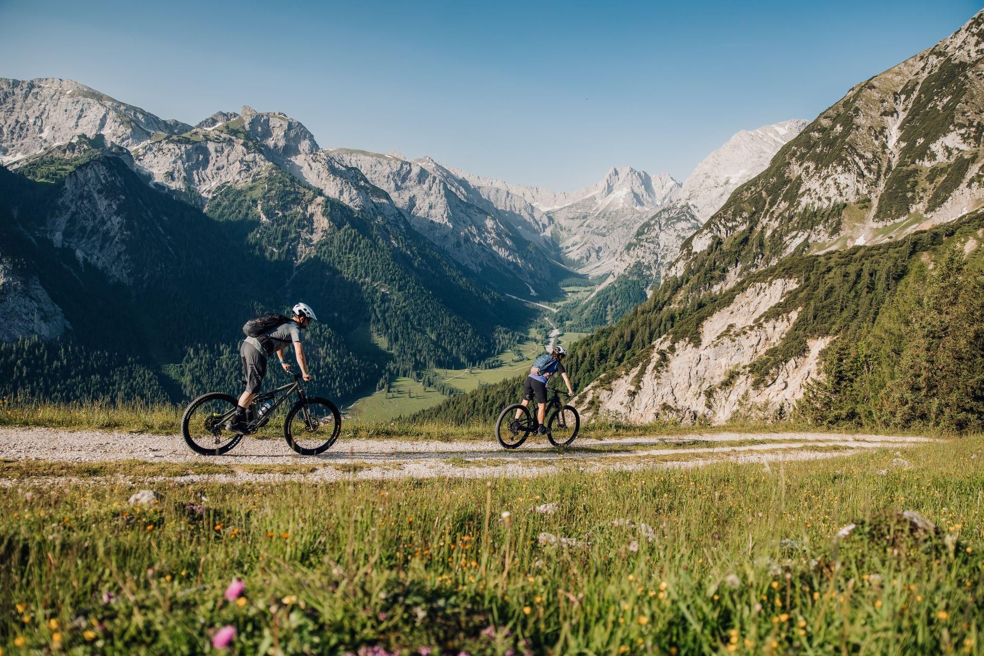 Zwei Radfahrer fahren auf einem schmalen Weg durch eine alpine Landschaft. Im Hintergrund sind hohe Berge und ein klarer Himmel zu sehen.