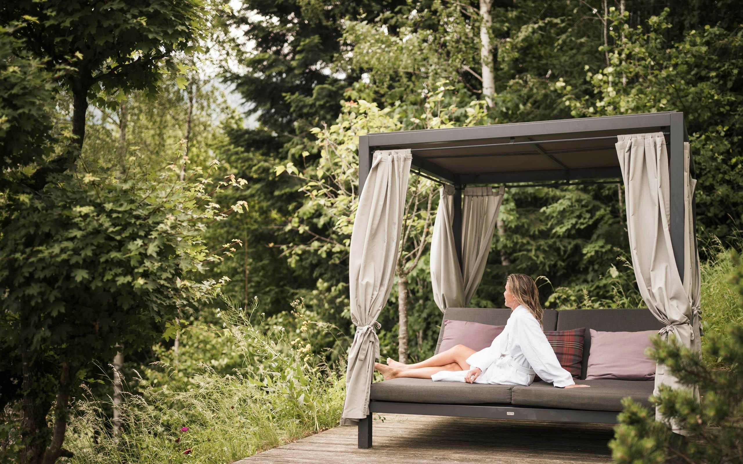 Eine Frau sitzt entspannt in einem gemütlichen Pavillon inmitten von grünem Gras und Bäumen. Der Ort strahlt Ruhe und Naturverbundenheit aus.