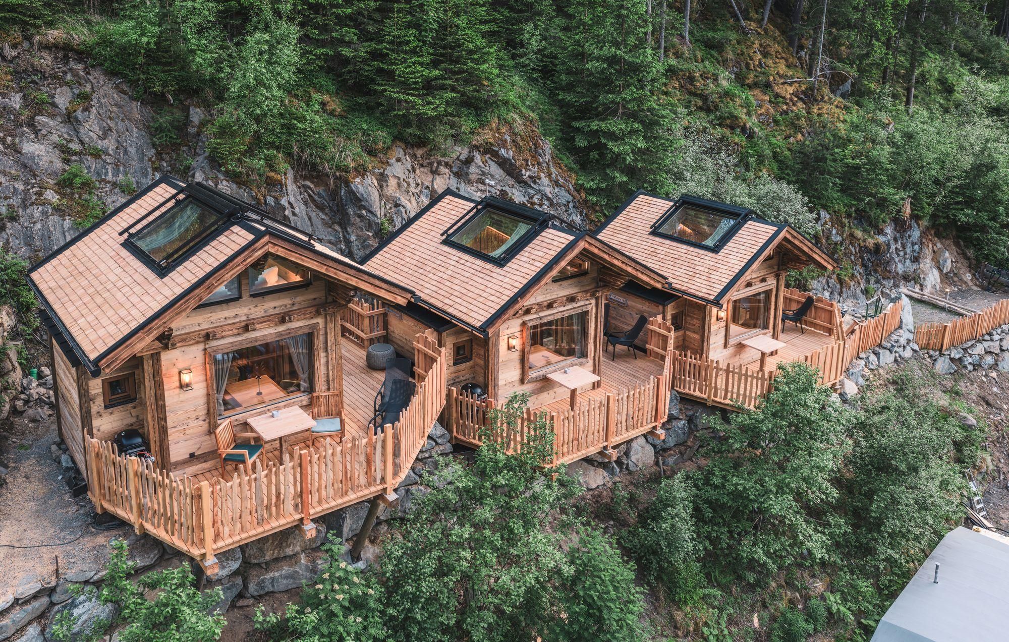 Drei Holzhäuschen stehen in der Natur, umgeben von Bäumen. Jedes Haus hat eine Veranda mit Ausblick auf die Umgebung.
