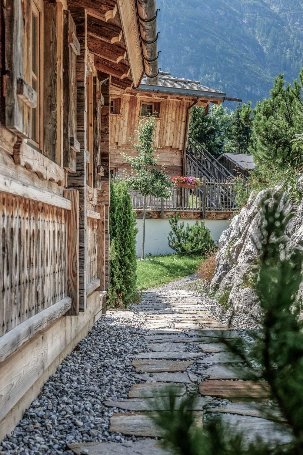 Ein malerischer Weg aus Steinen führt zwischen Chalets vorbei. Umgeben von grünen Pflanzen und Bergen vermittelt die Szene eine ruhige Atmosphäre.