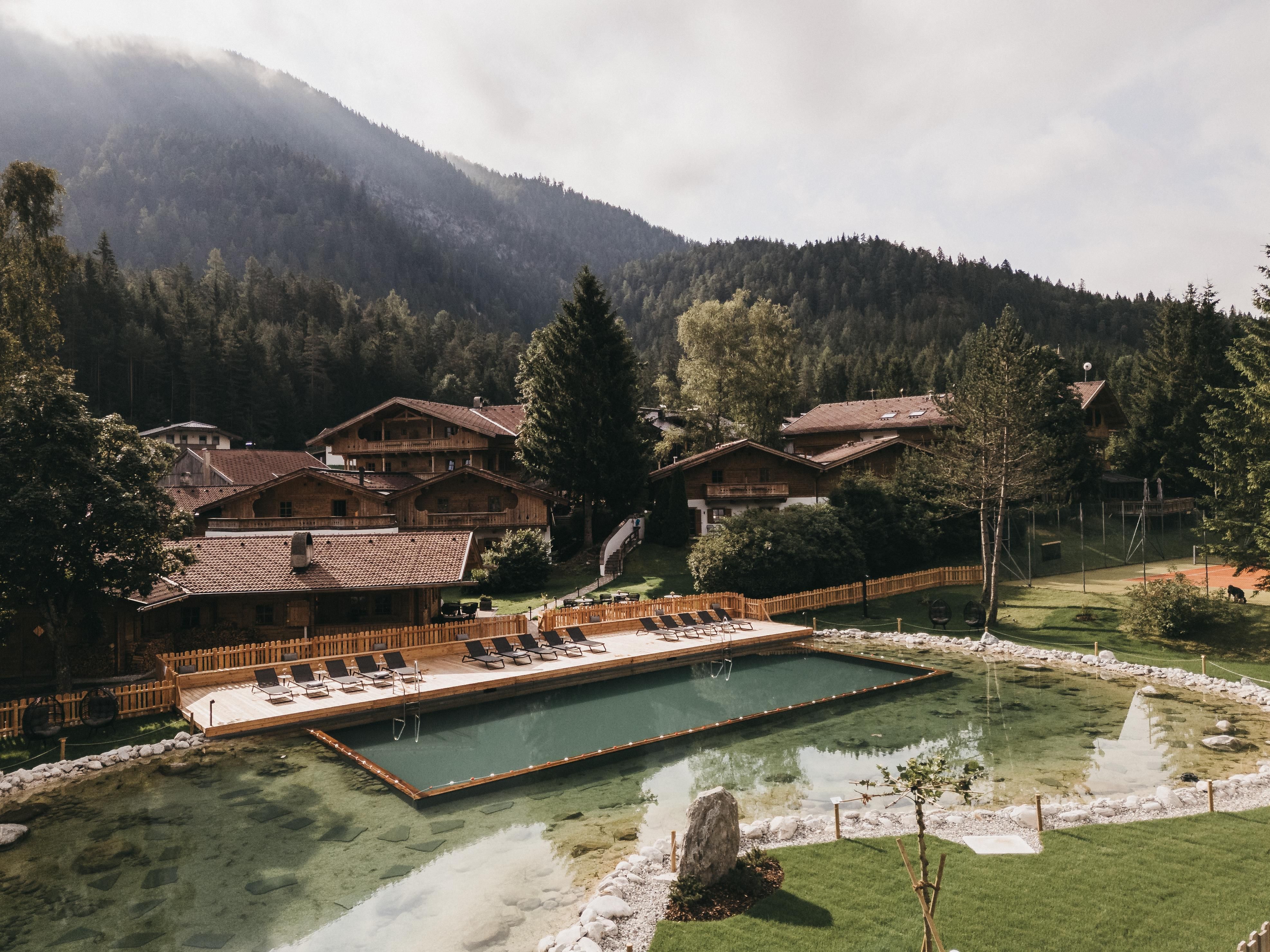 Ein malerisches Bergland mit einem kleinen Schwimmteich und gemütlichen Holzchalets. Die Umgebung ist von Bäumen und Bergen umgeben, was eine entspannte Atmosphäre schafft.