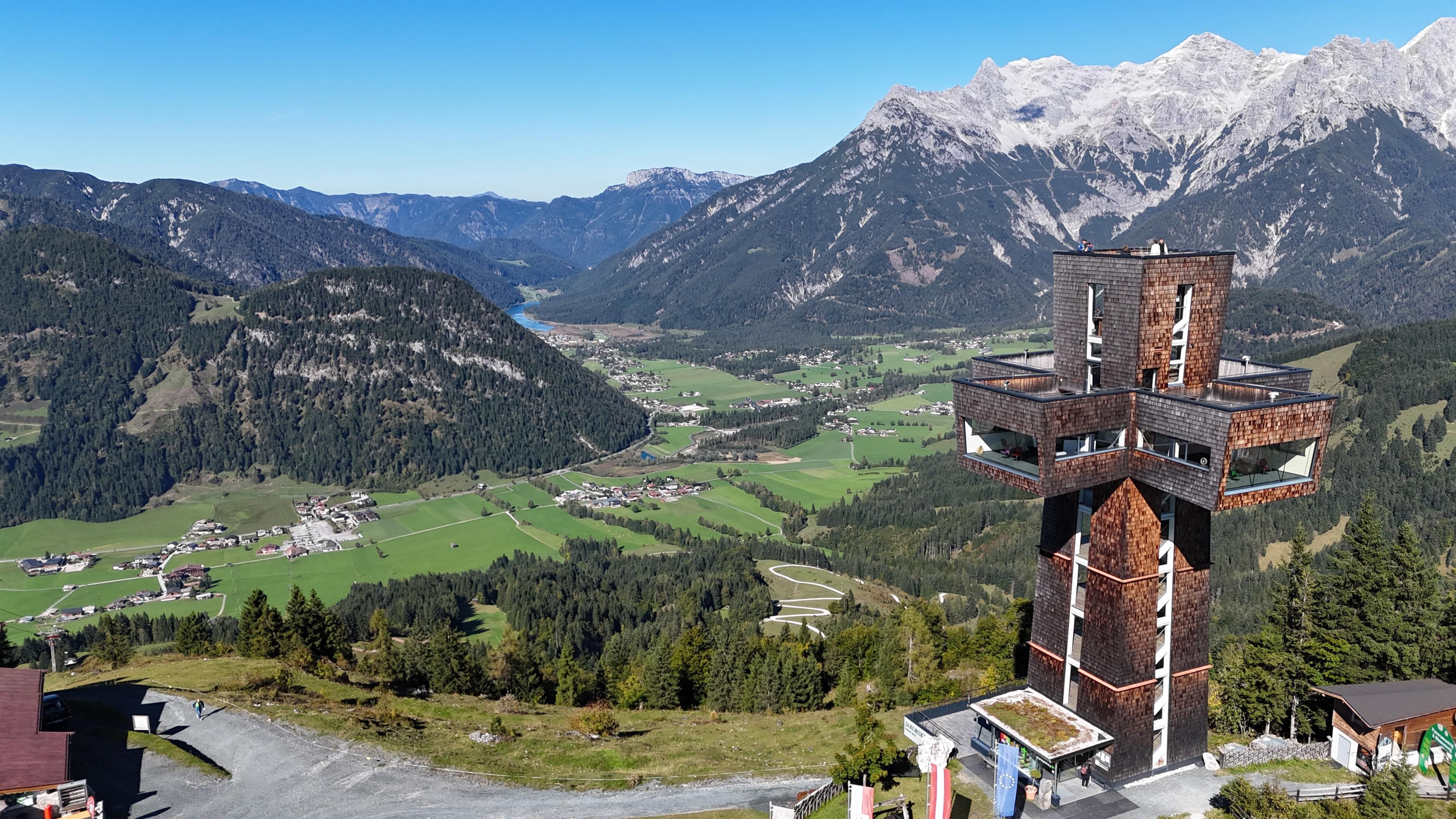 Eine beeindruckende Berglandschaft mit einem Aussichtsturm im Vordergrund. Im Hintergrund sind schneebedeckte Berge und grüne Täler zu sehen.